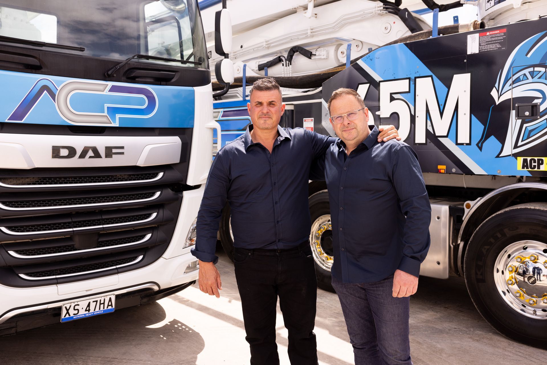 Two men stand in front of two concrete mixer trucks, posing. One man has his arm around the other. Trucks have blue and white branding.