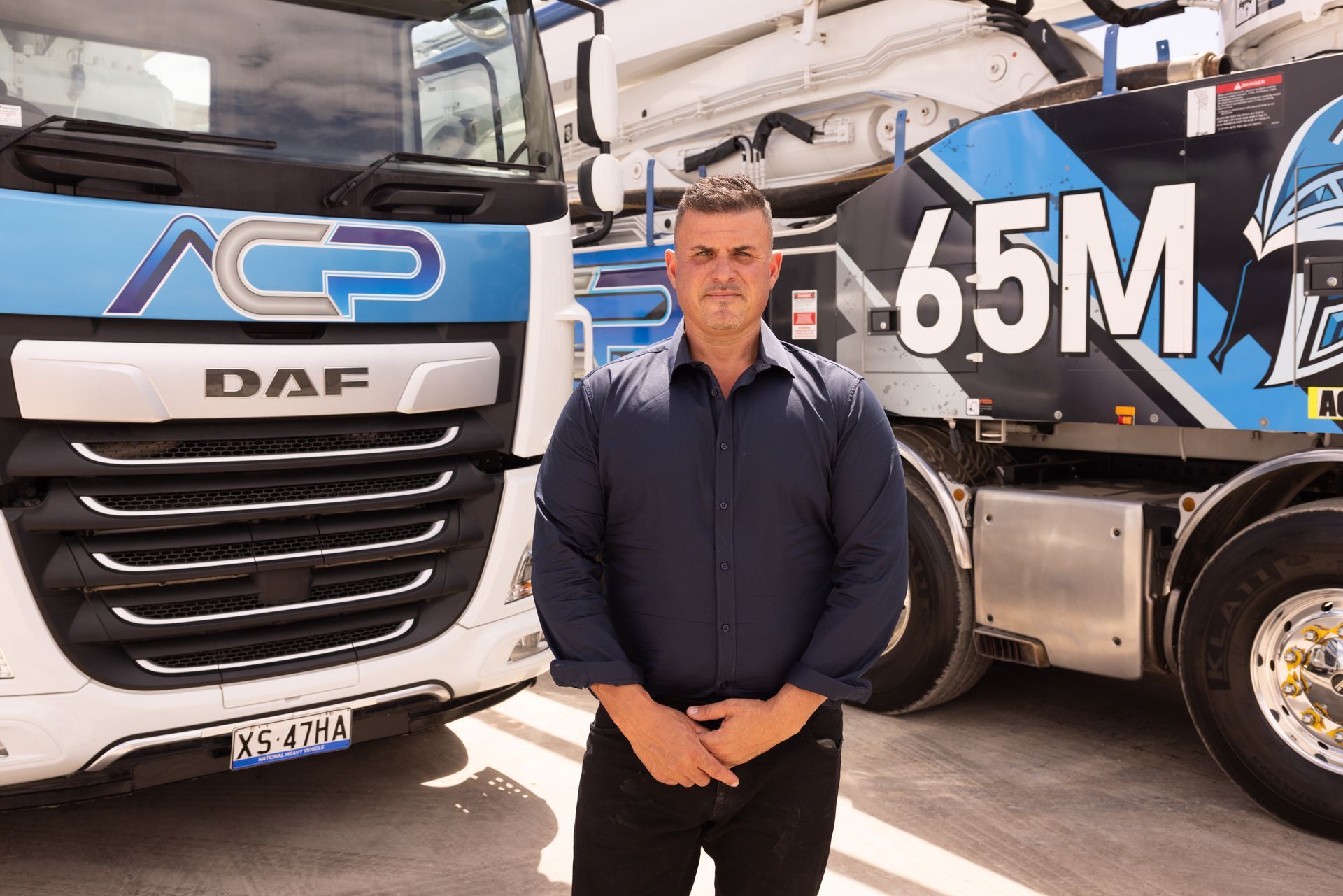 Man standing in front of a concrete truck. He's wearing a blue shirt and black pants. Trucks have blue and white markings.