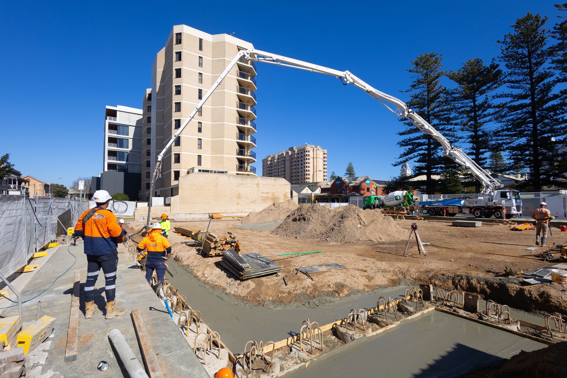 Construction site: concrete pouring with pump truck; workers, building under construction. Sunny day.