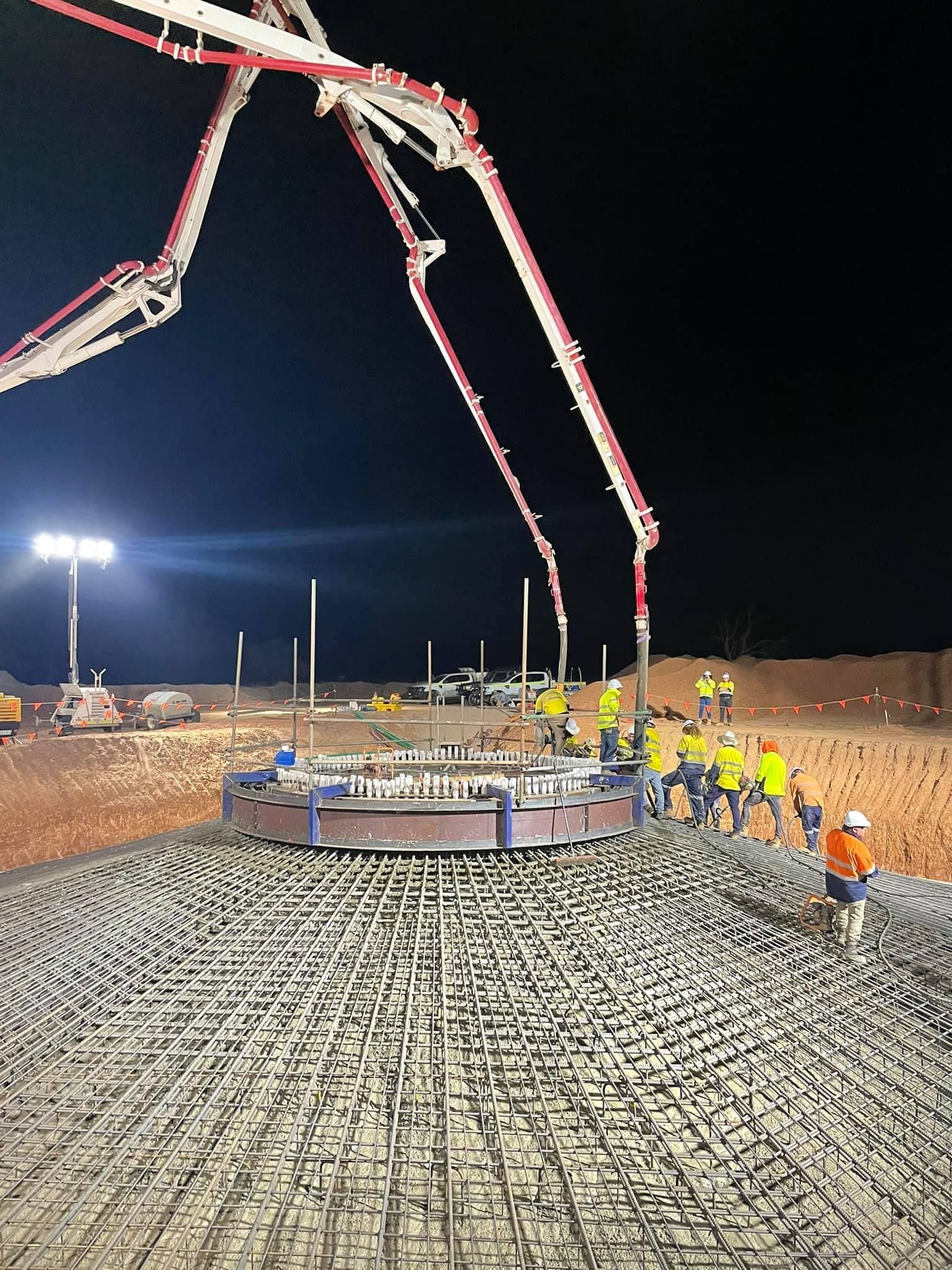 Concrete pour at night: Concrete pump reaching over rebar-covered structure with workers in hard hats.