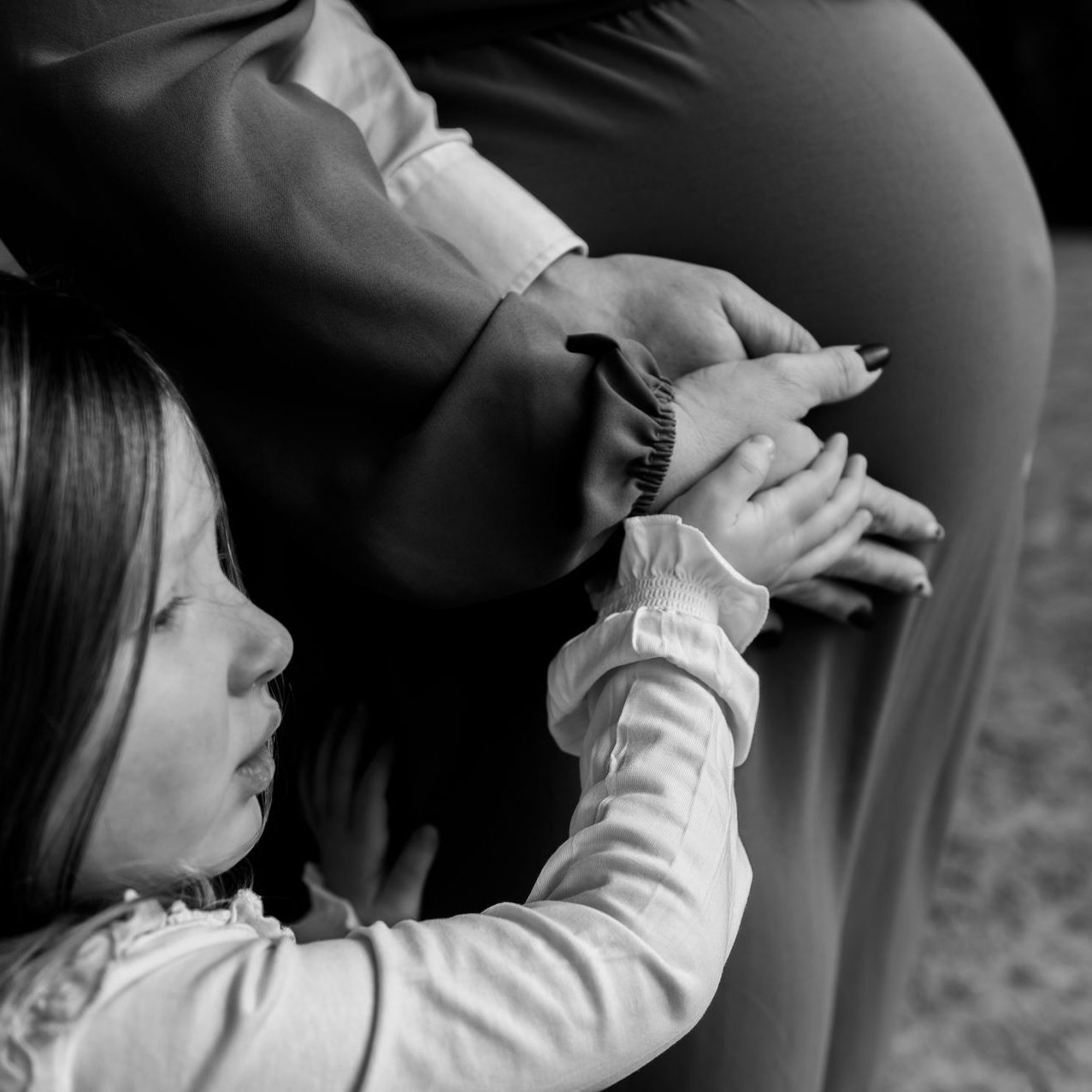 De buik van een zwangere vrouw met een klein kindje dat ertegenaan ligt, binnenshuis, zwart-wit.