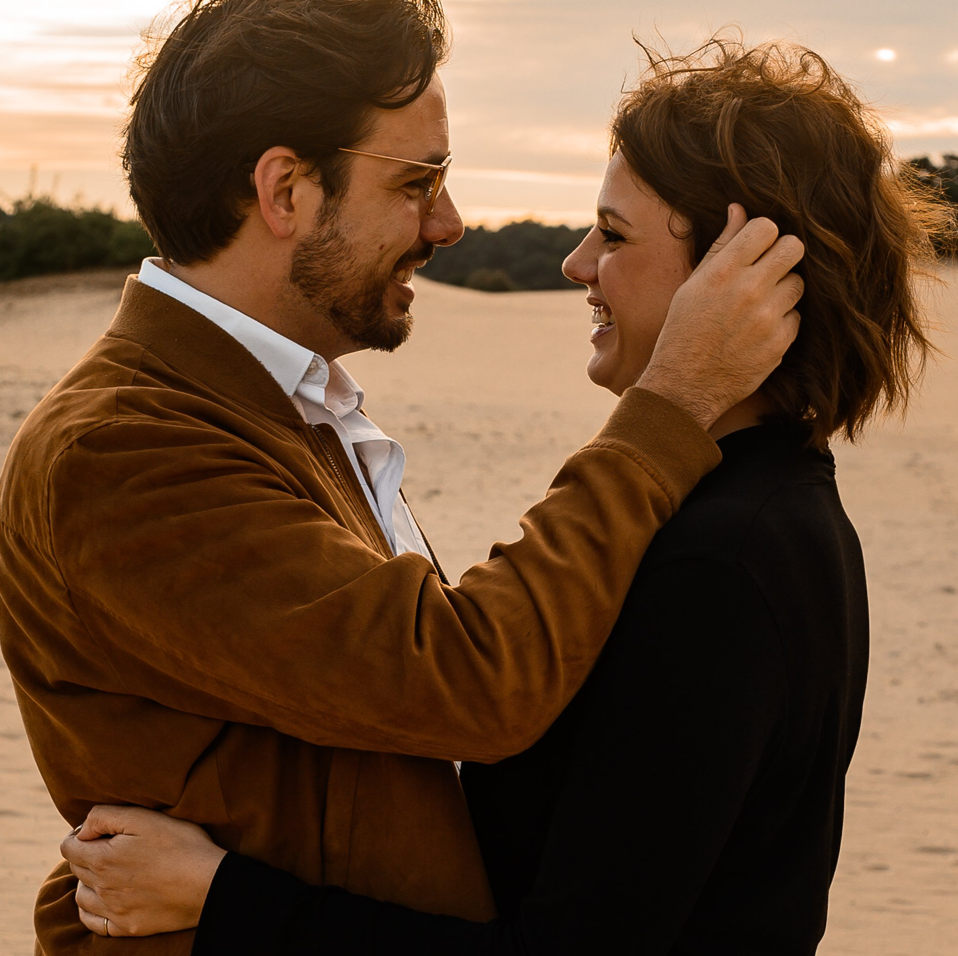 Een man in een bruine jas omhelst een vrouw in een zwarte top in de buitenlucht, terwijl ze elkaar glimlachend aankijken.