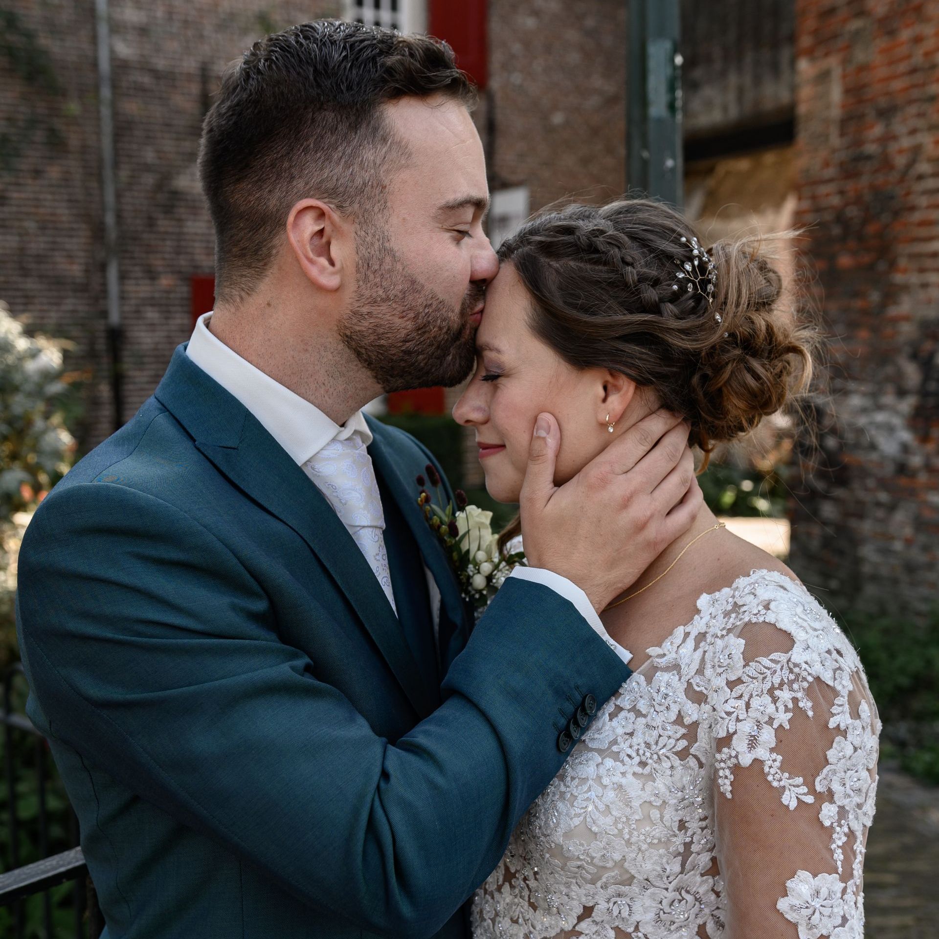 De bruidegom kust de bruid op haar voorhoofd en houdt haar gezicht teder vast. De bruiloft vindt plaats in een bakstenen gebouw.
