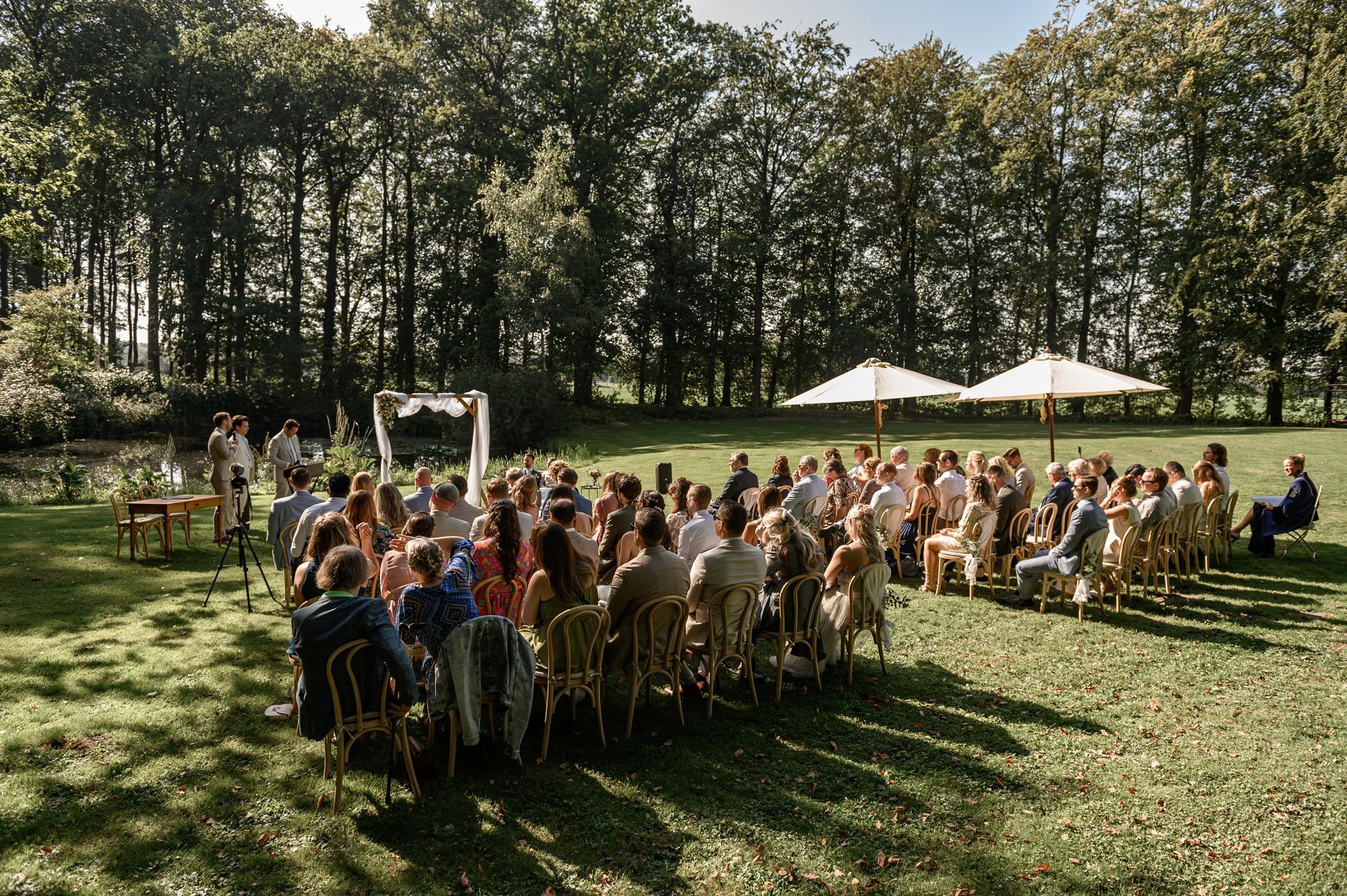 Een grote groep mensen zit op stoelen in een grasveld tijdens een huwelijksceremonie.