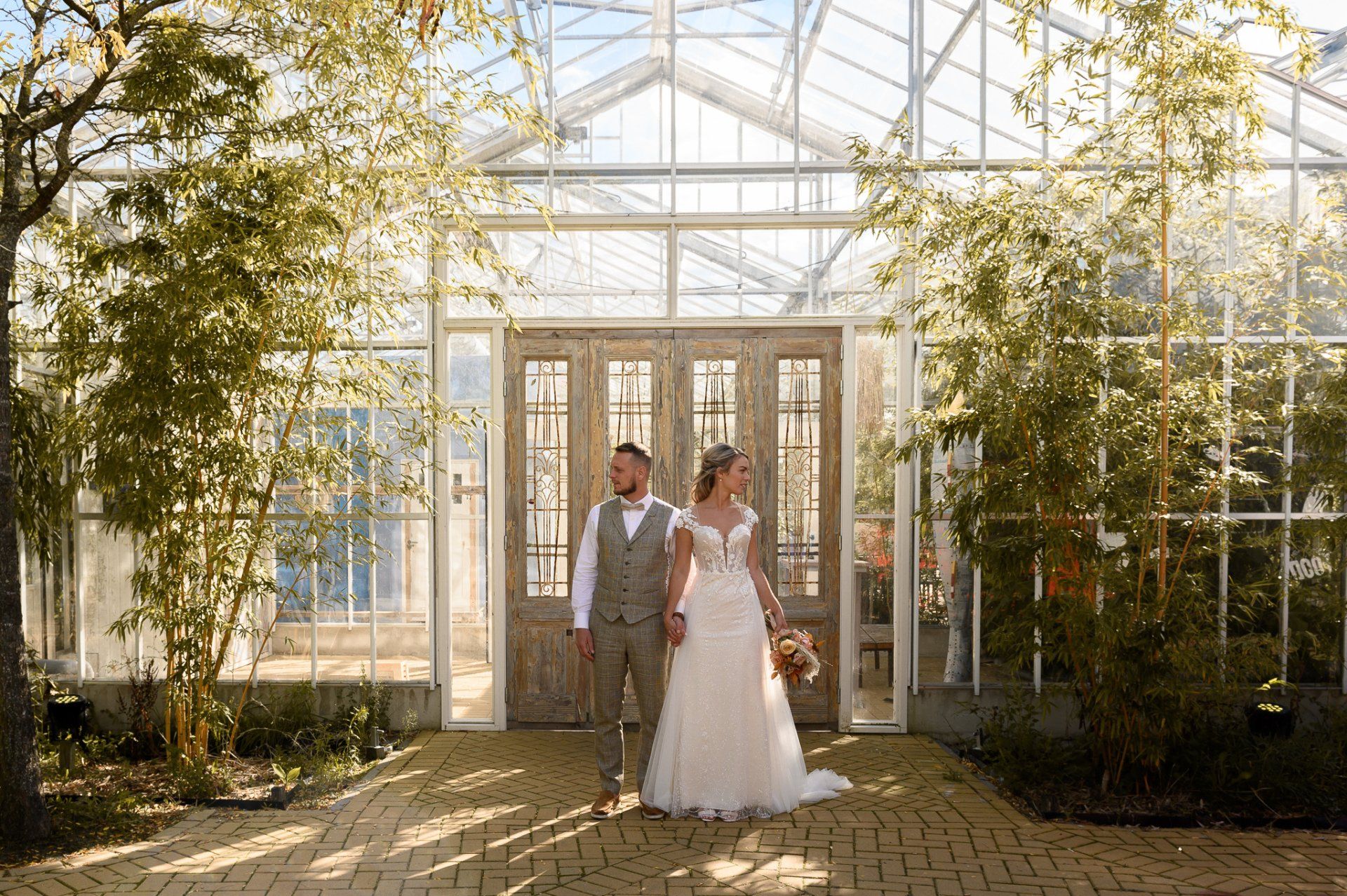 Een bruidspaar staat hand in hand voor een kas.