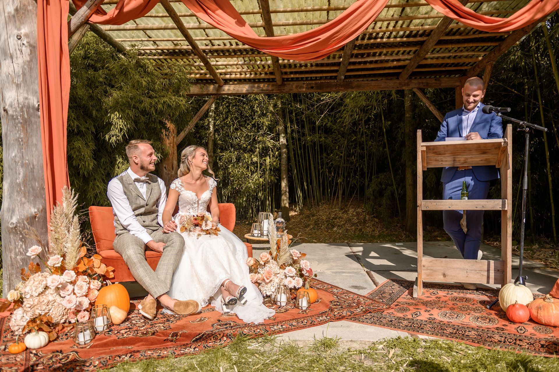 Een bruid en bruidegom zitten op een bank, terwijl een man een toespraak houdt.