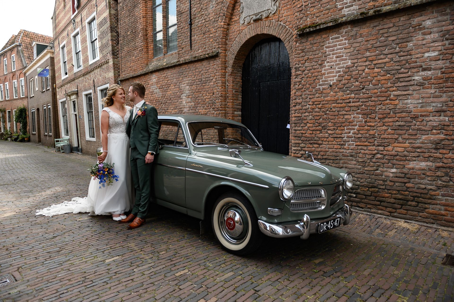 Een bruid en bruidegom staan naast een groene auto tijdens de geposeerde shoot.