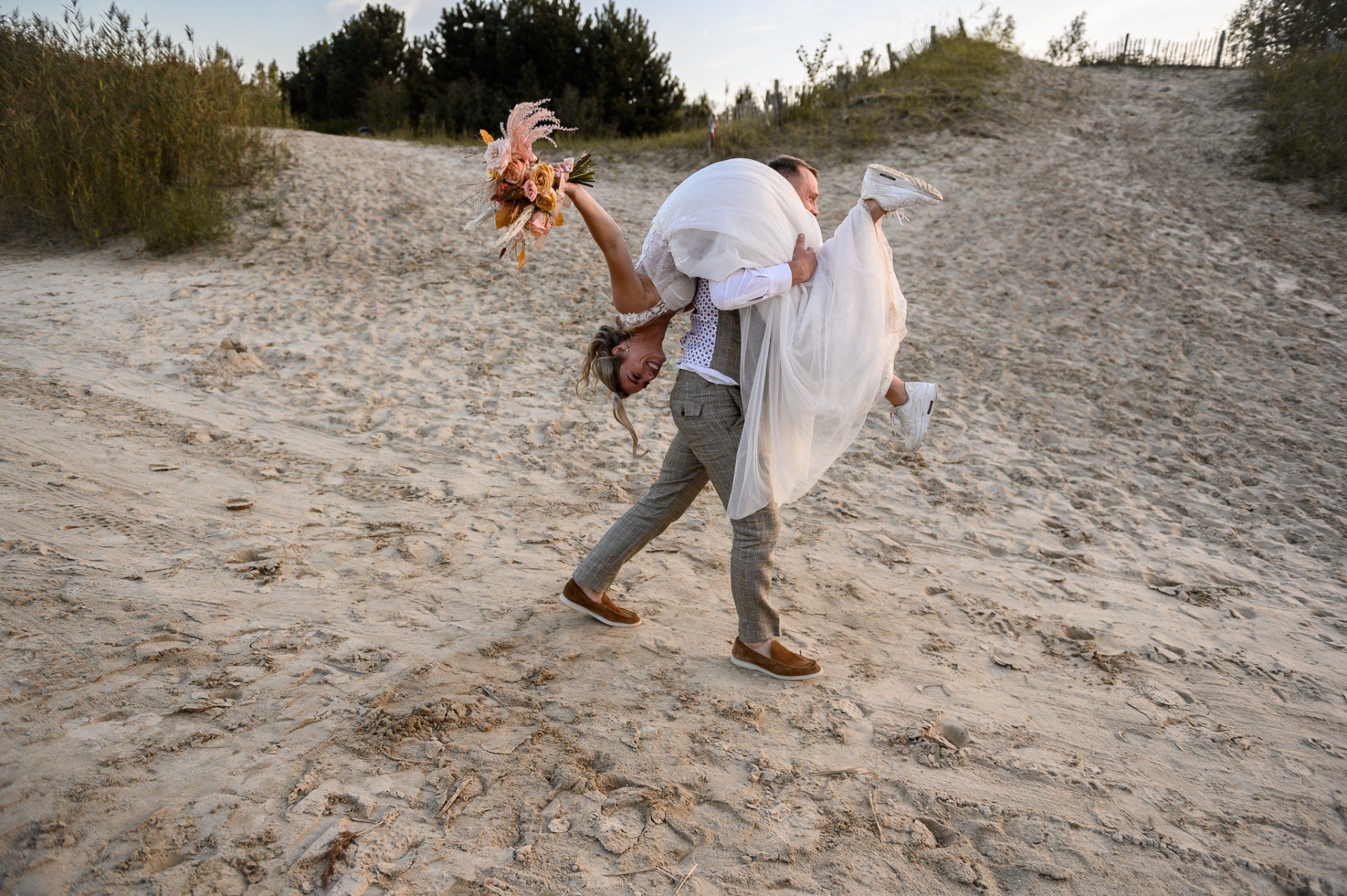 Een man draagt een bruid op zijn schouders in het zand.