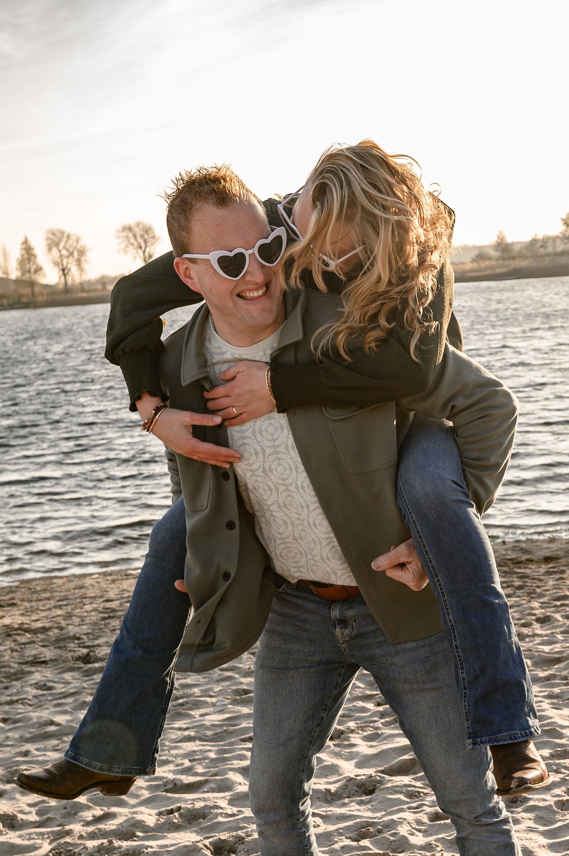 Een man geeft een vrouw een ritje op zijn rug op het strand; beiden dragen een zonnebril en glimlachen.