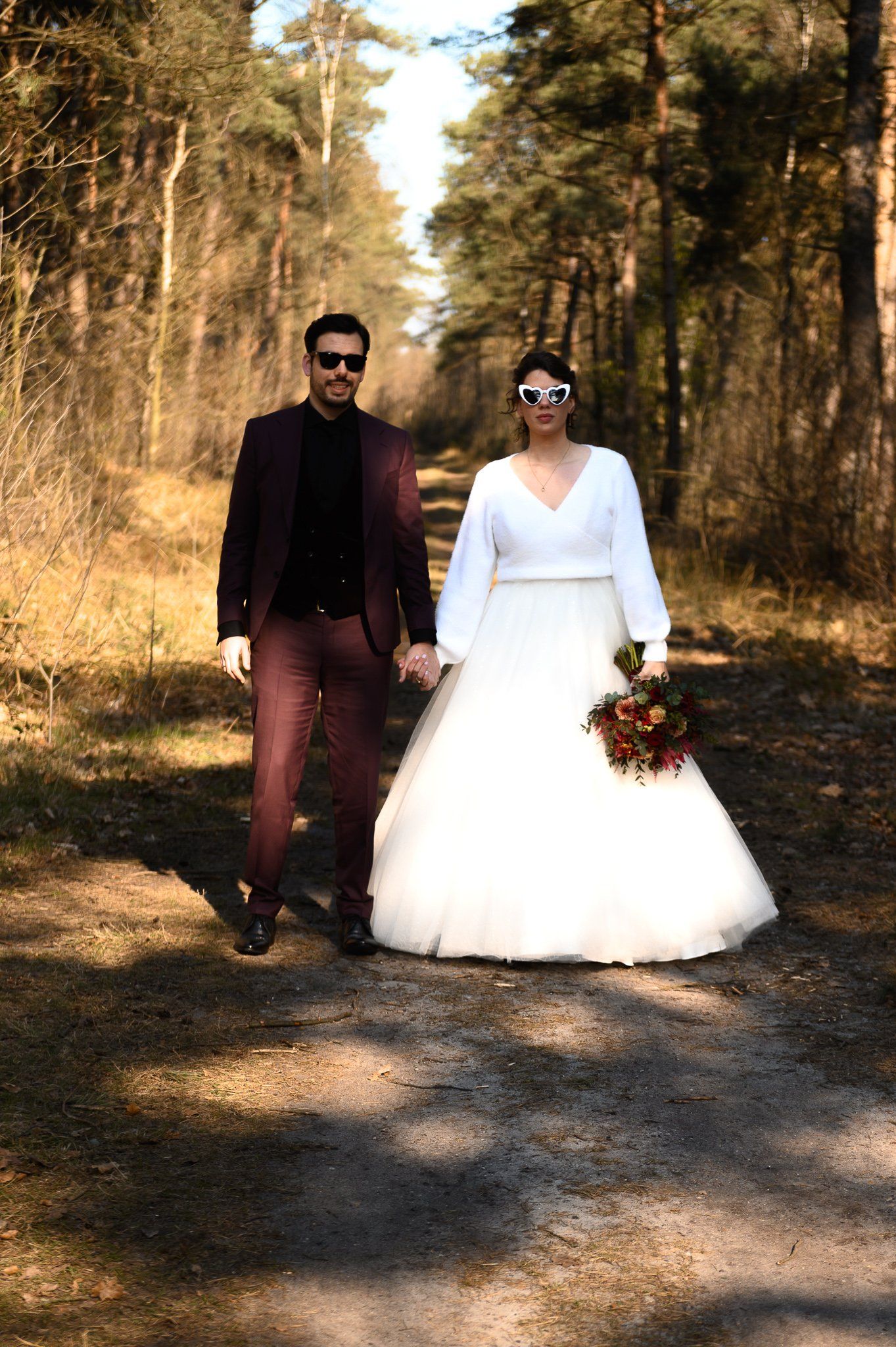 Een bruid en bruidegom lopen hand in hand over een pad in het bos.