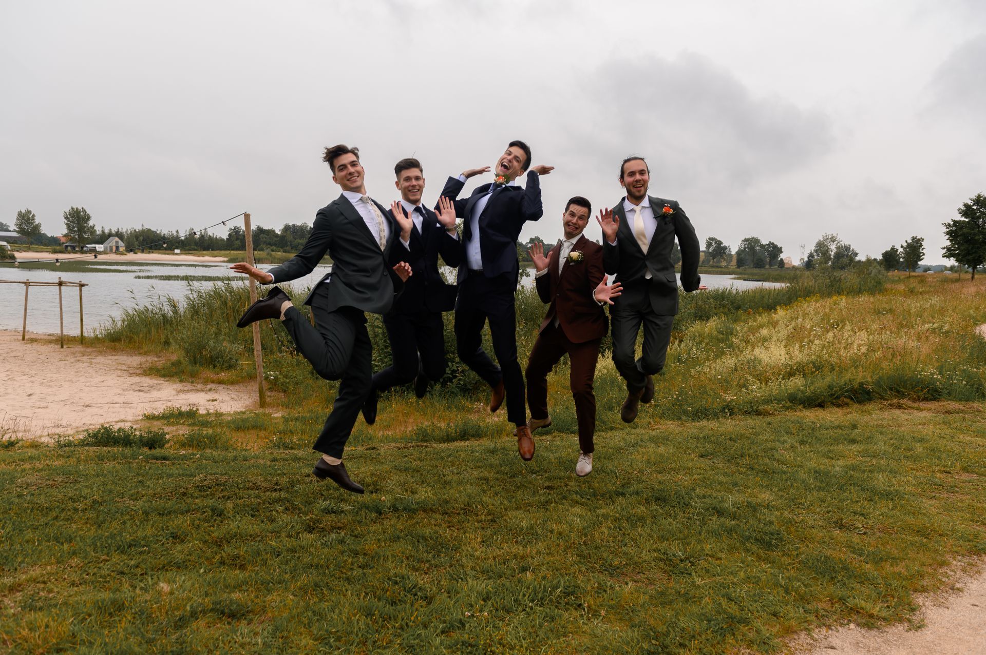 Een groep vrienden in pakken springt in de lucht in een veld samen met bruidegom.