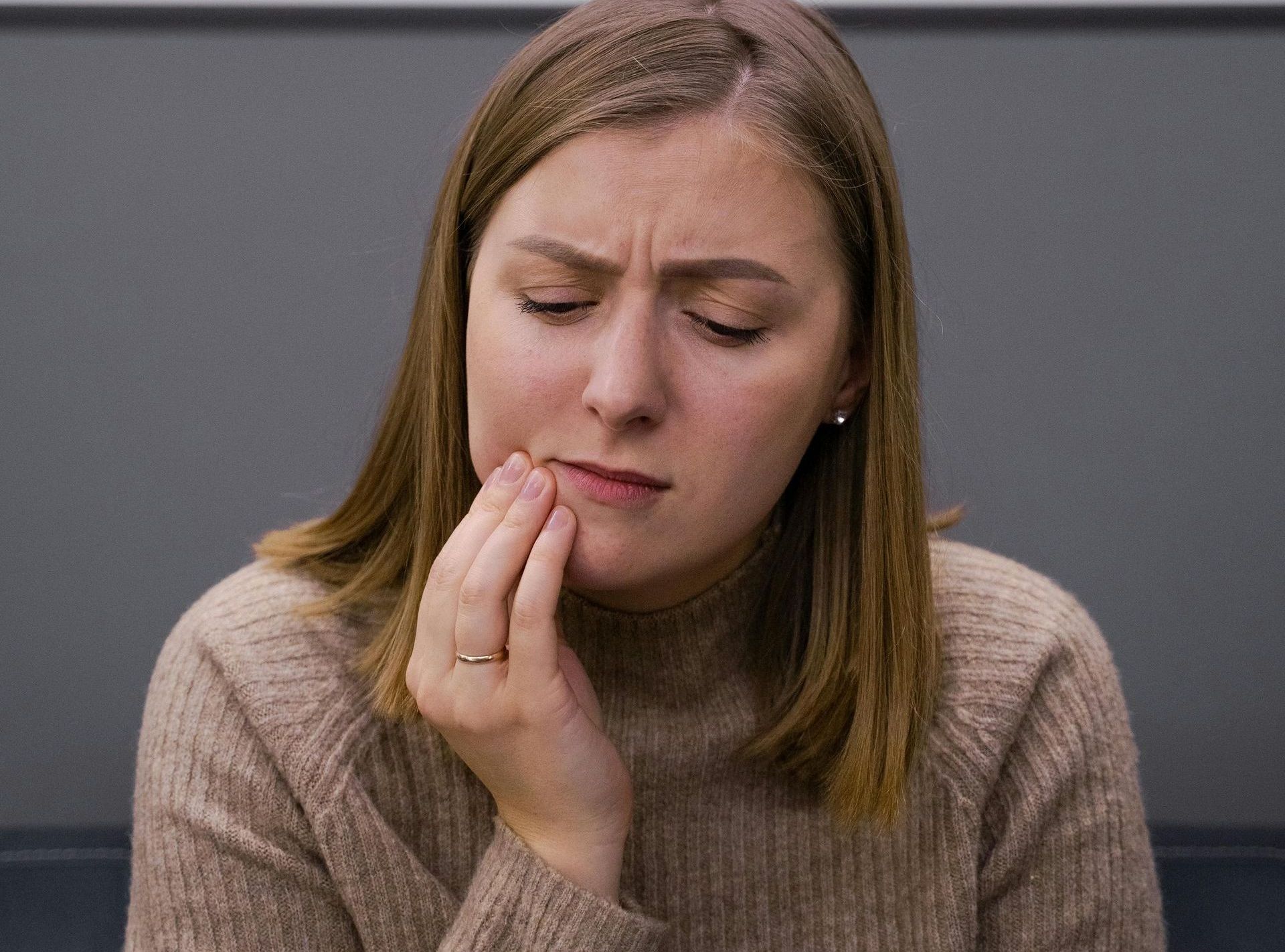 A woman in a brown sweater is holding her mouth in pain.