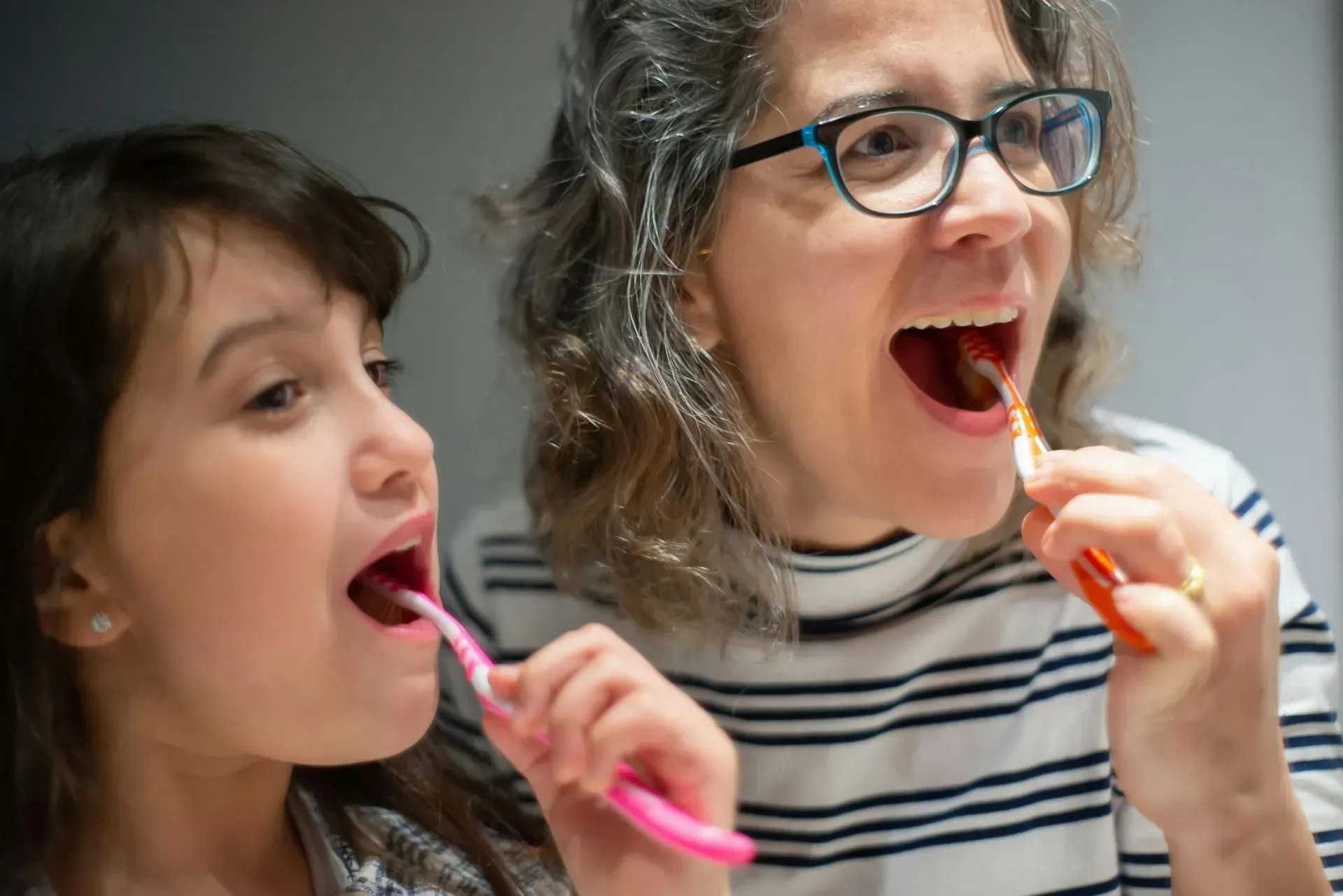 A woman and a little girl are brushing their teeth together in front of a mirror.