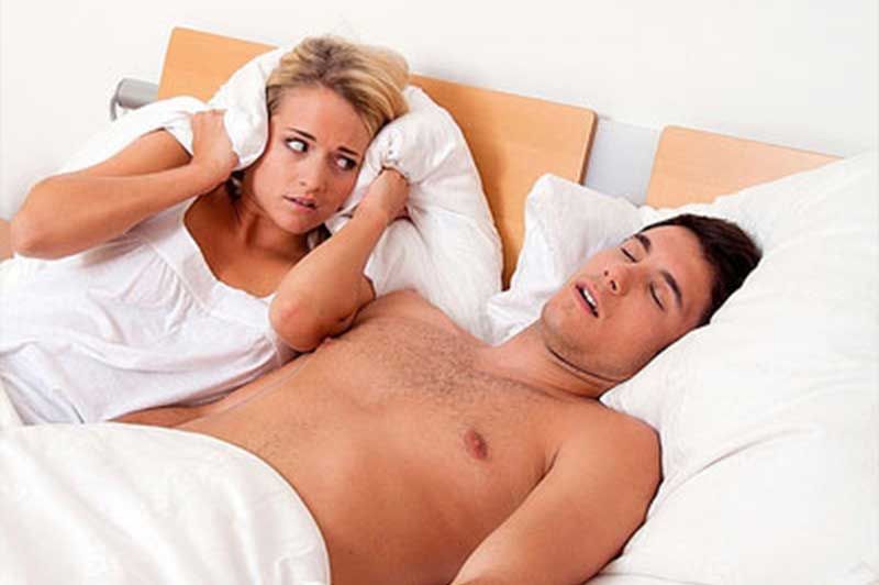 A woman is covering her ears while a man sleeps in bed.