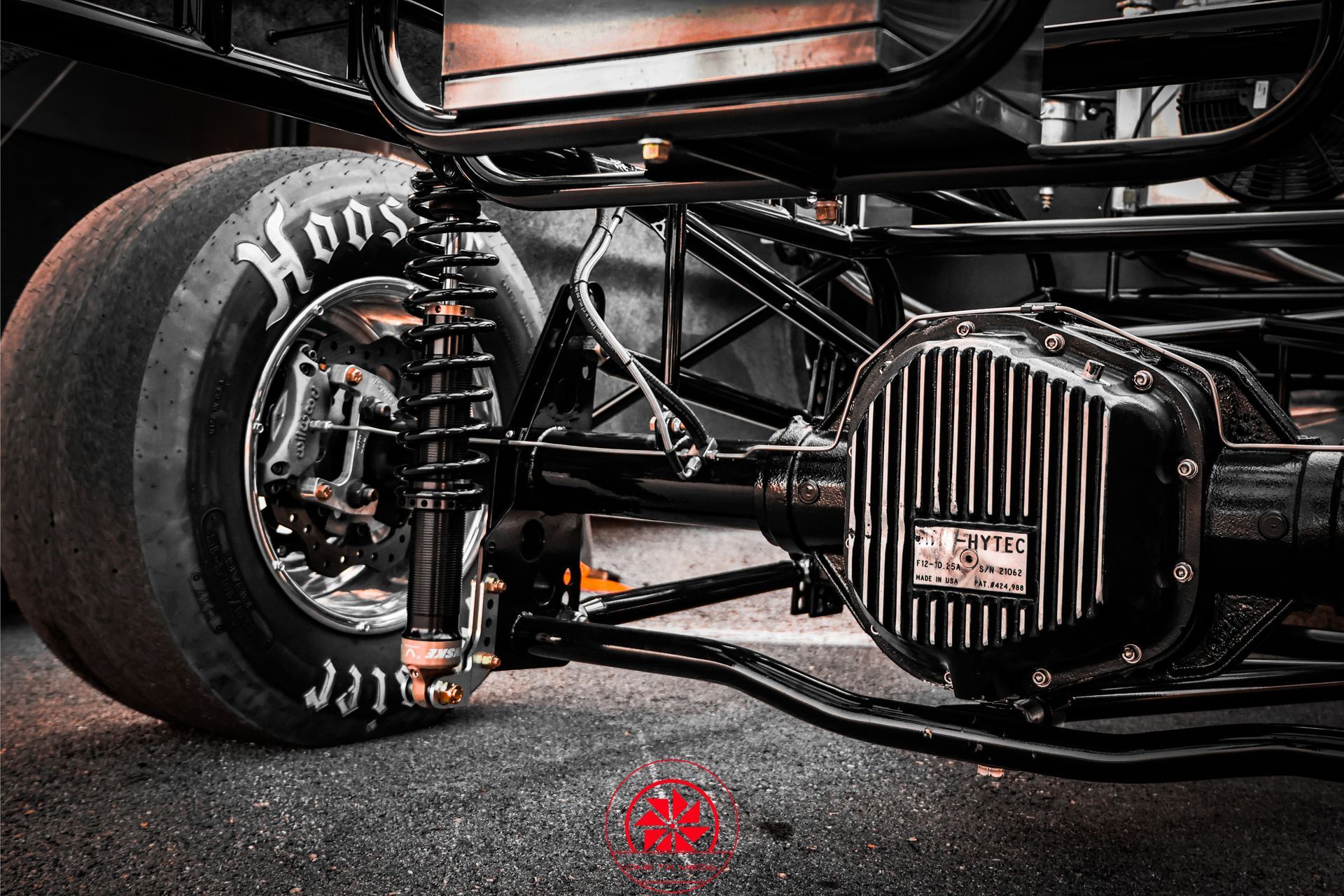 Close-up of a black dragster’s front suspension and tire on a racetrack, with exposed engine components.