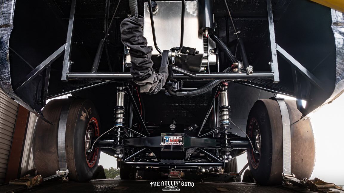 Rear view of a black race car with wide tires and suspension undercarriage on a track at dusk