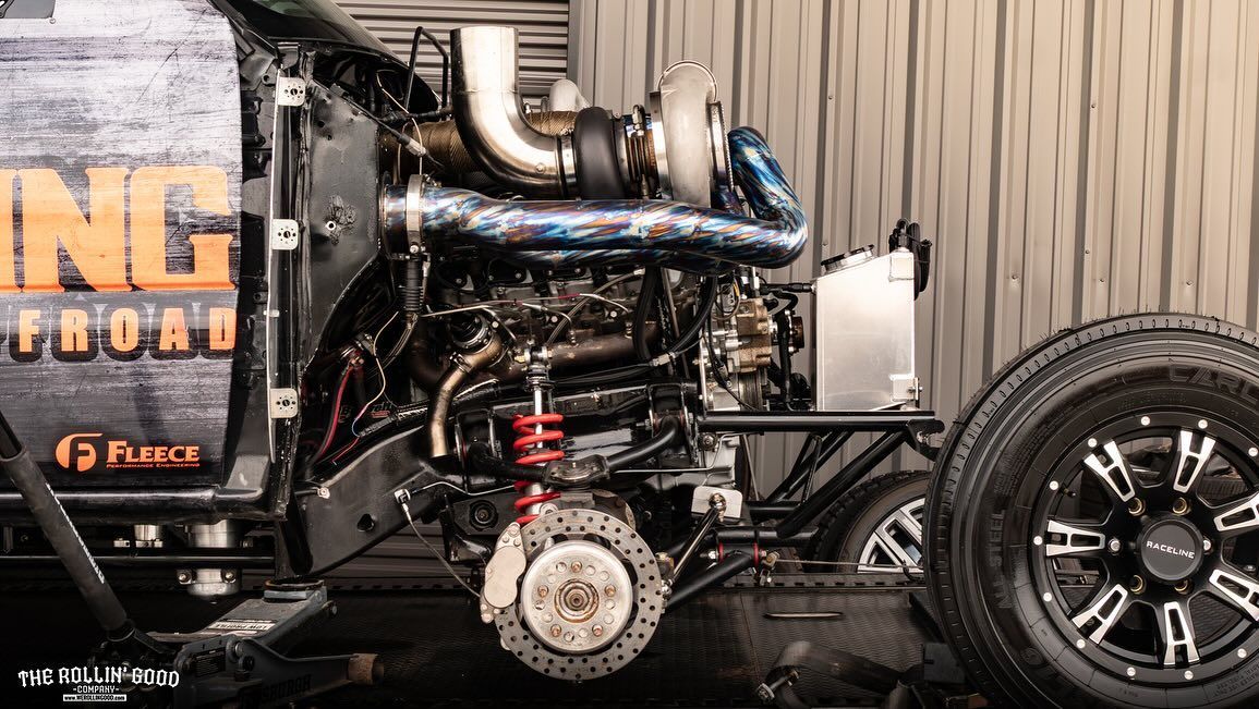 Black custom hot rod engine and front wheel at a car show, with orange lettering on the rear body panel