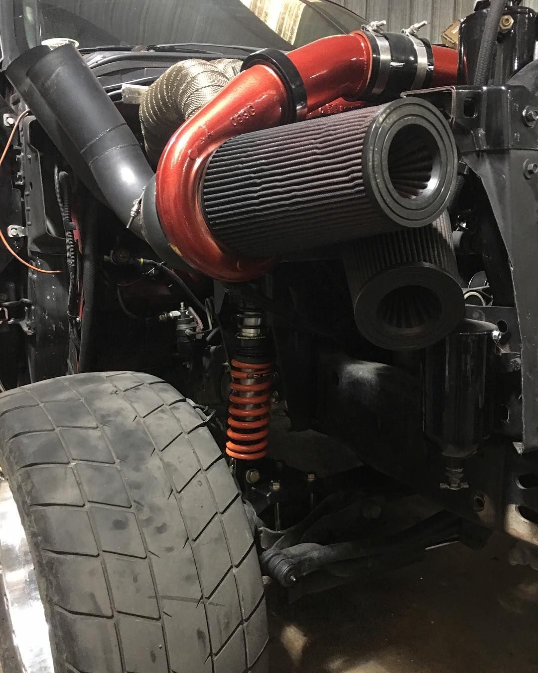 Close-up of a race car’s front suspension, large tire, and red intake piping with black filters.