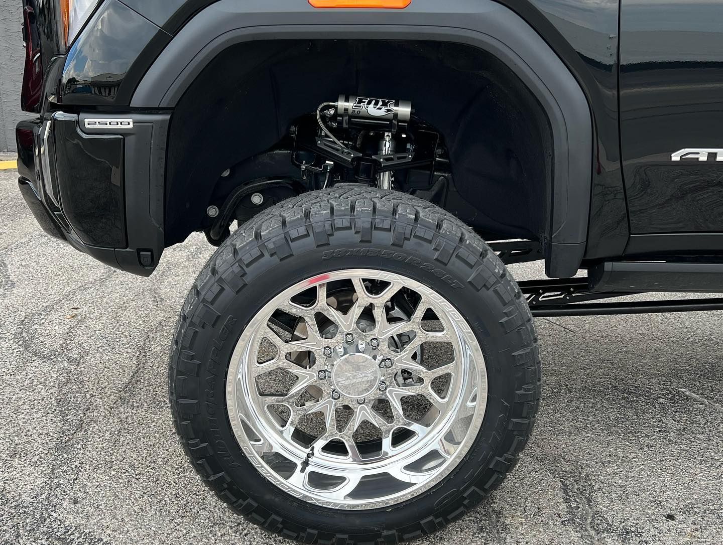 Black truck with large chrome wheel and off-road tire parked on gravel.
