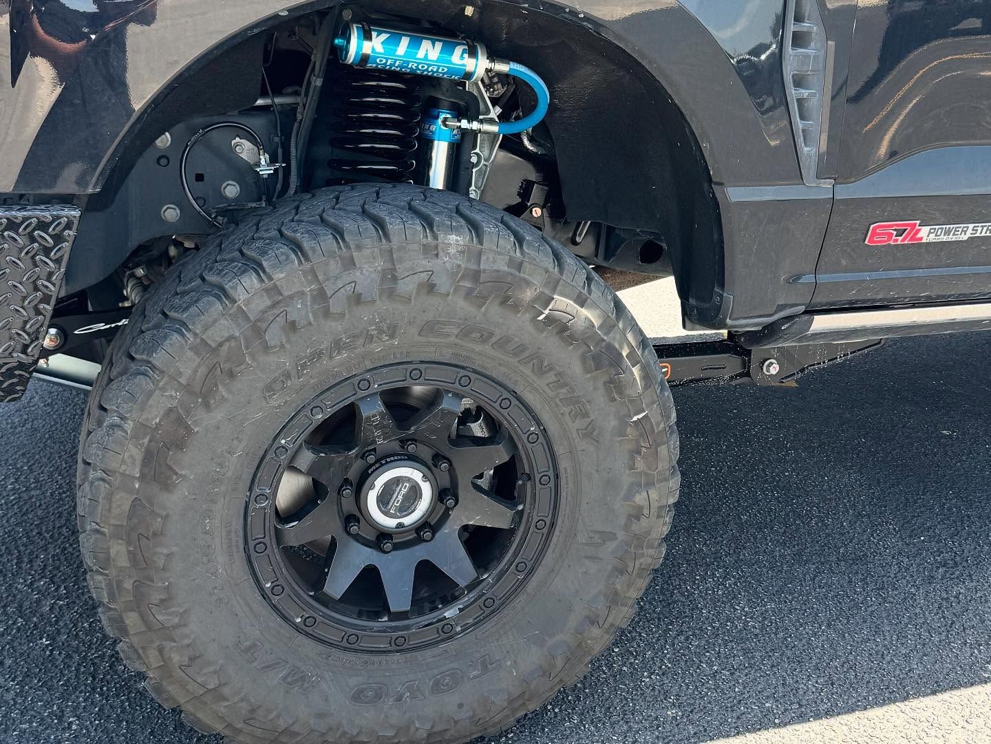 Close-up of a black off-road truck’s front wheel and suspension on a paved road.