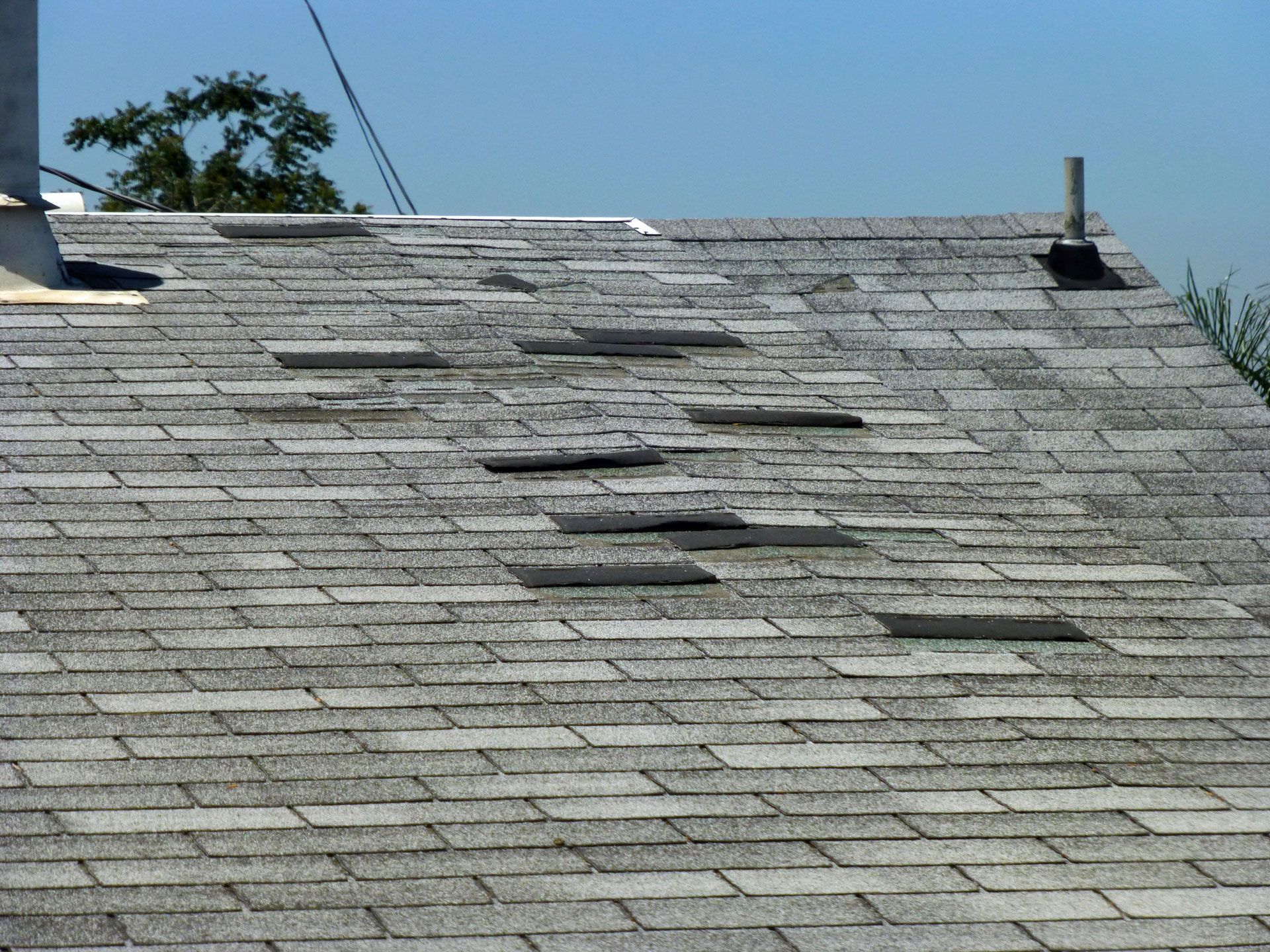 Damaged gray asphalt shingle roof with missing and broken shingles on a sunny day.
