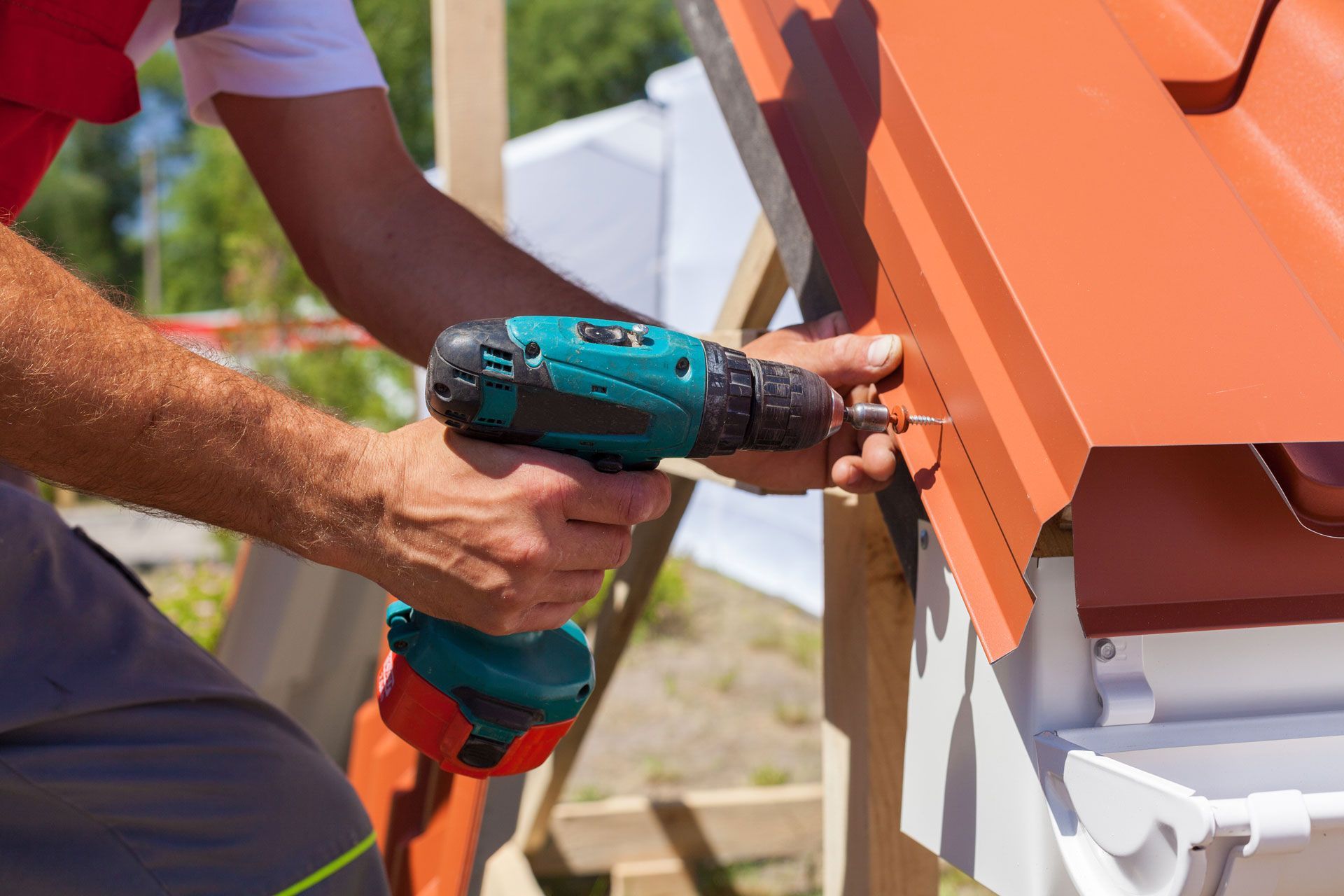 Person using a power drill to install a red metal roof panel, outdoors.