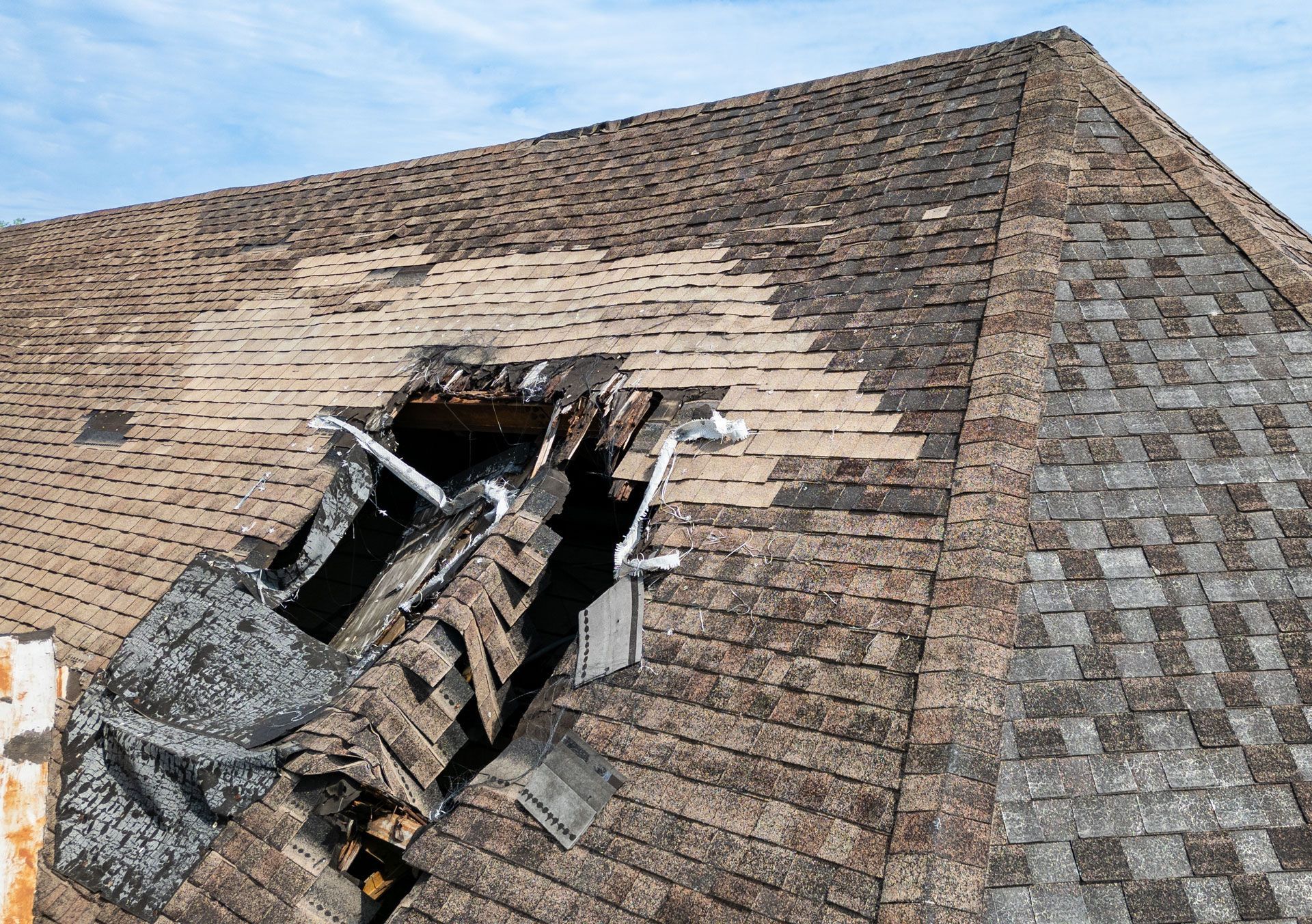Damaged asphalt shingle roof with a large hole. Shingles are brown and weathered, with visible underlayment.