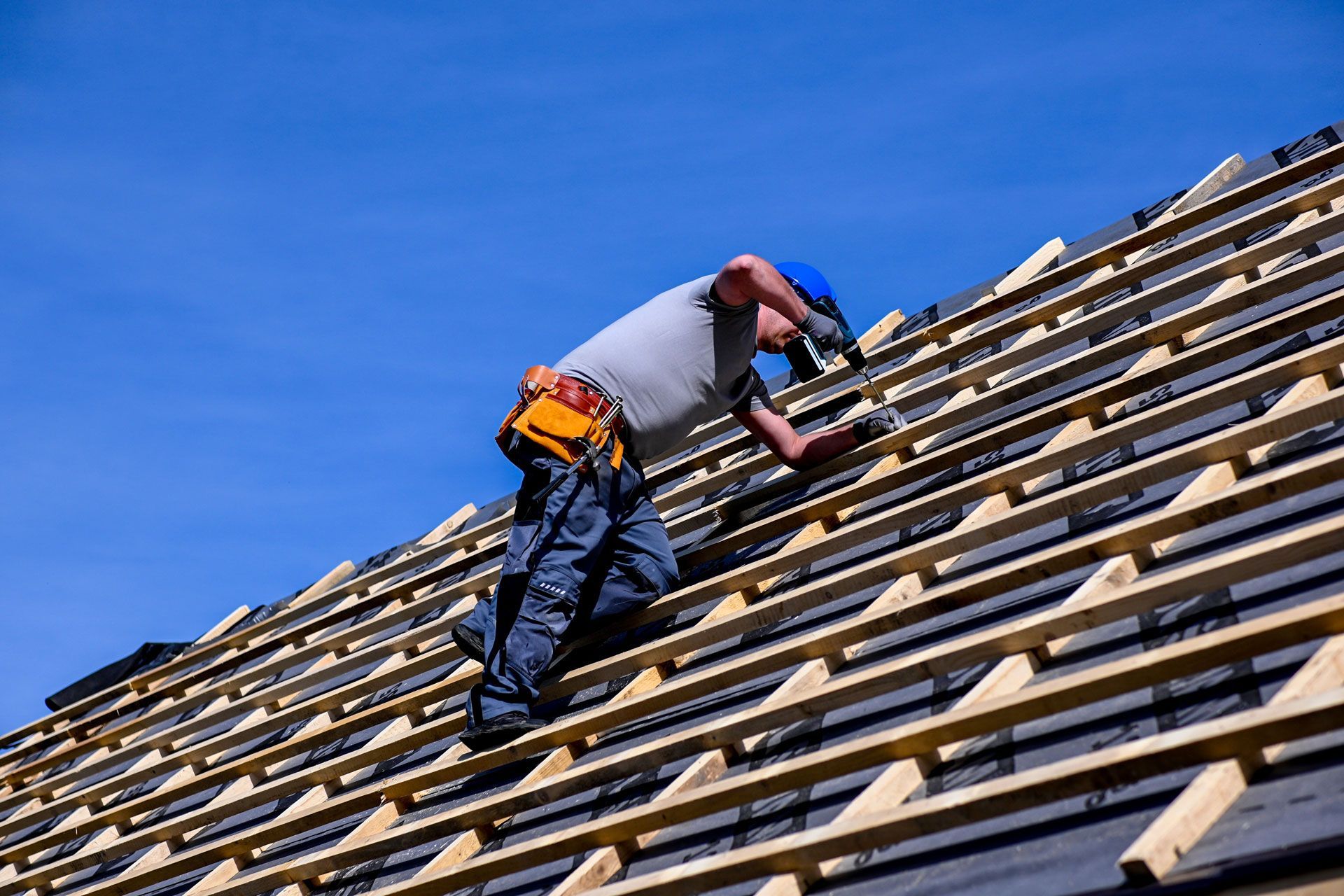 Roofer installing shingles on a roof under a bright blue sky.