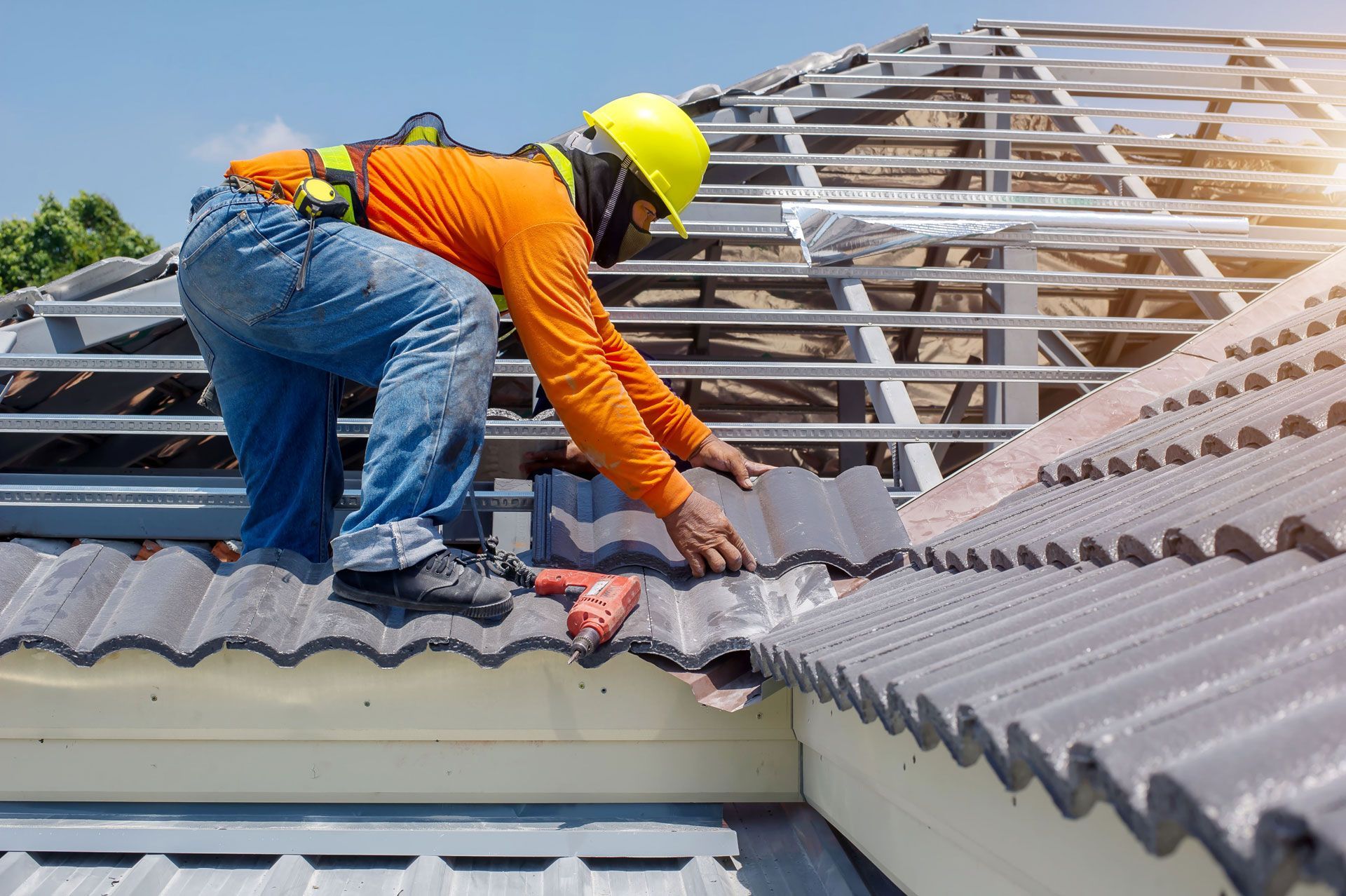 Roofer in safety gear installing gray tiles on a partially constructed roof. Bright blue sky.