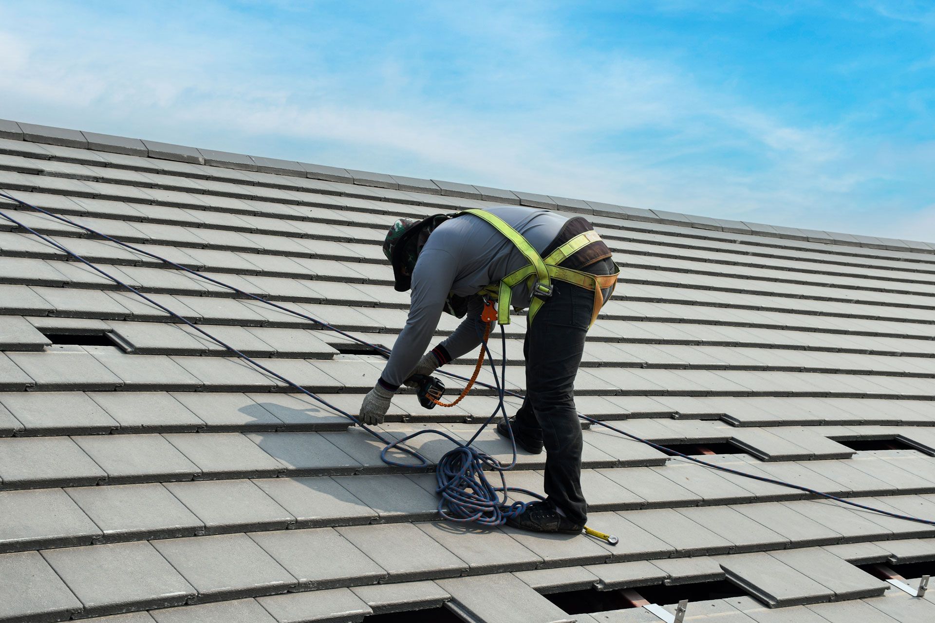 Roofer wearing safety harness working on gray tiled roof under a blue sky.