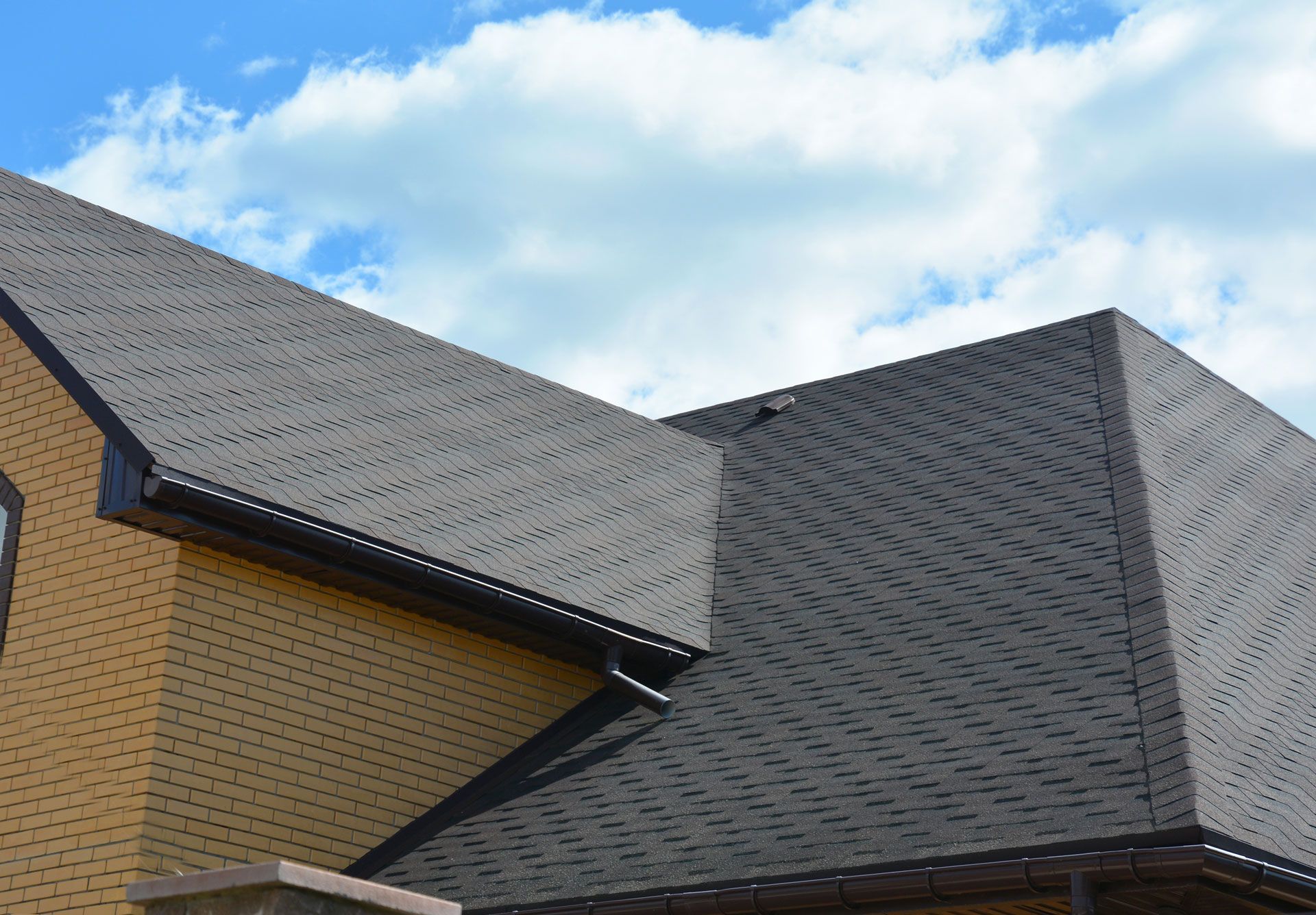 Brown brick house with dark gray shingle roof under a blue sky with fluffy white clouds.