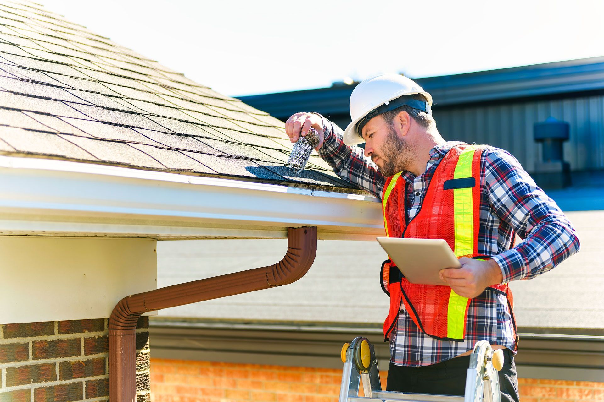 Man in hardhat and vest inspecting roof gutter, holding tablet.