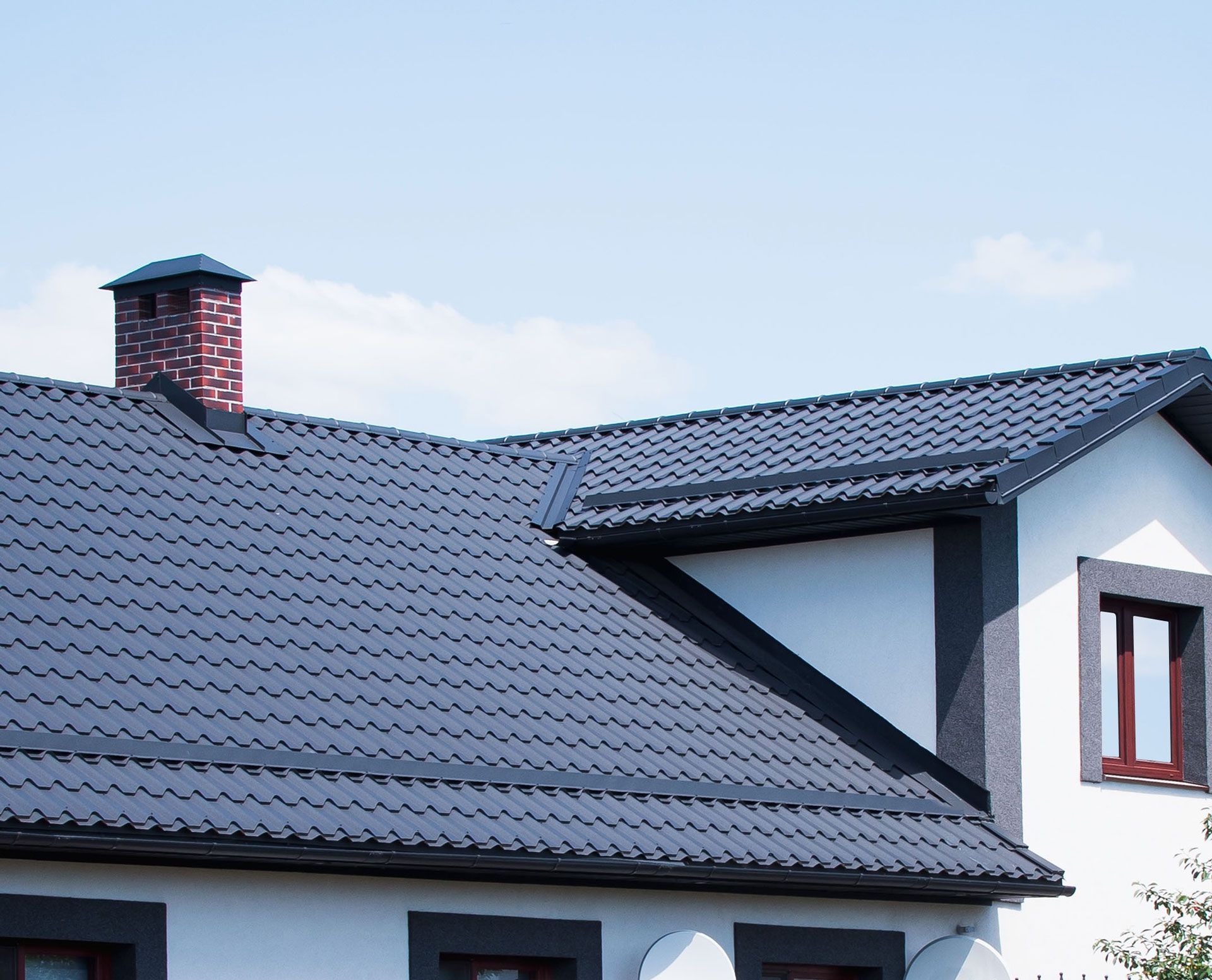 Grey tiled roof on a white house with a brick chimney against a blue sky.