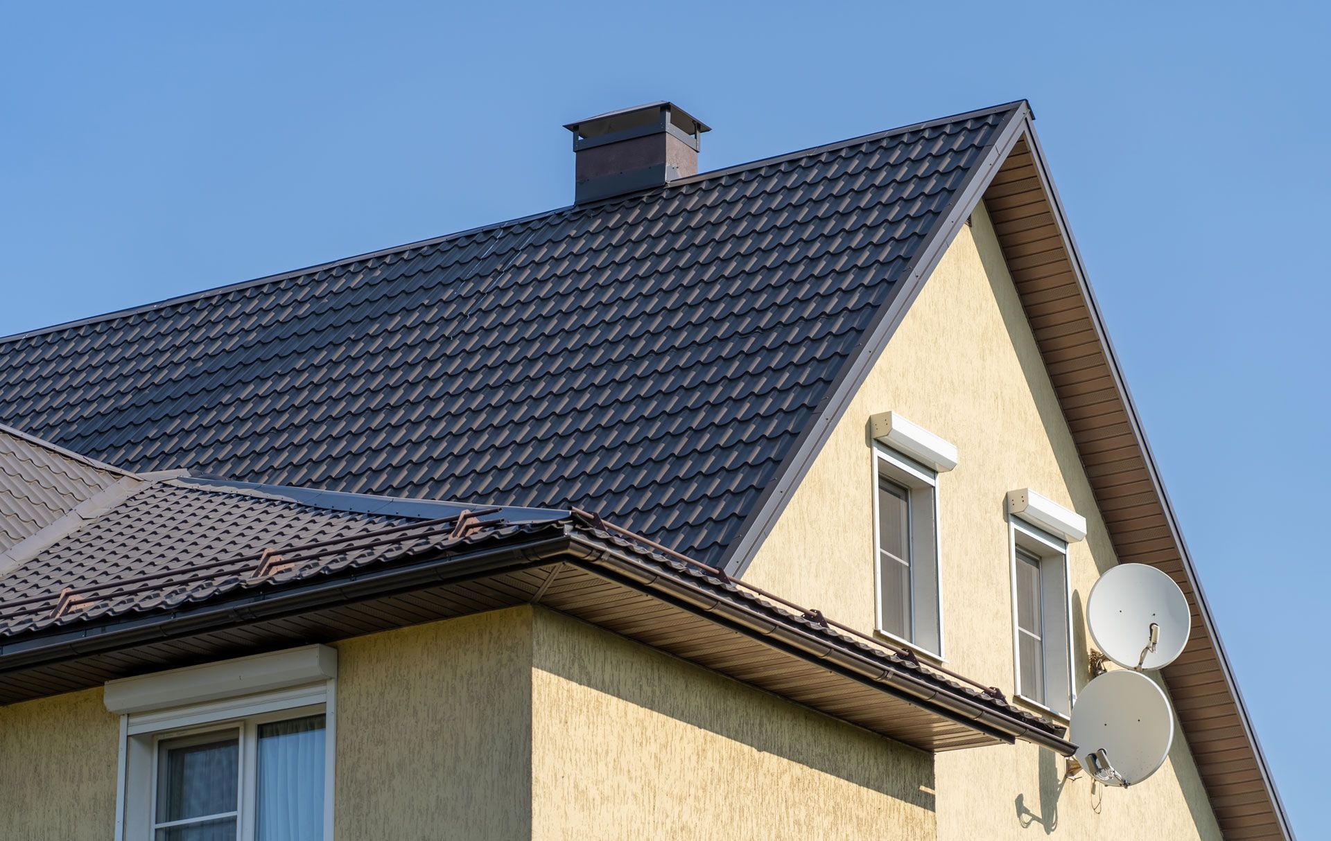 Yellow house with a dark gray roof, chimney, and satellite dishes against a clear blue sky.