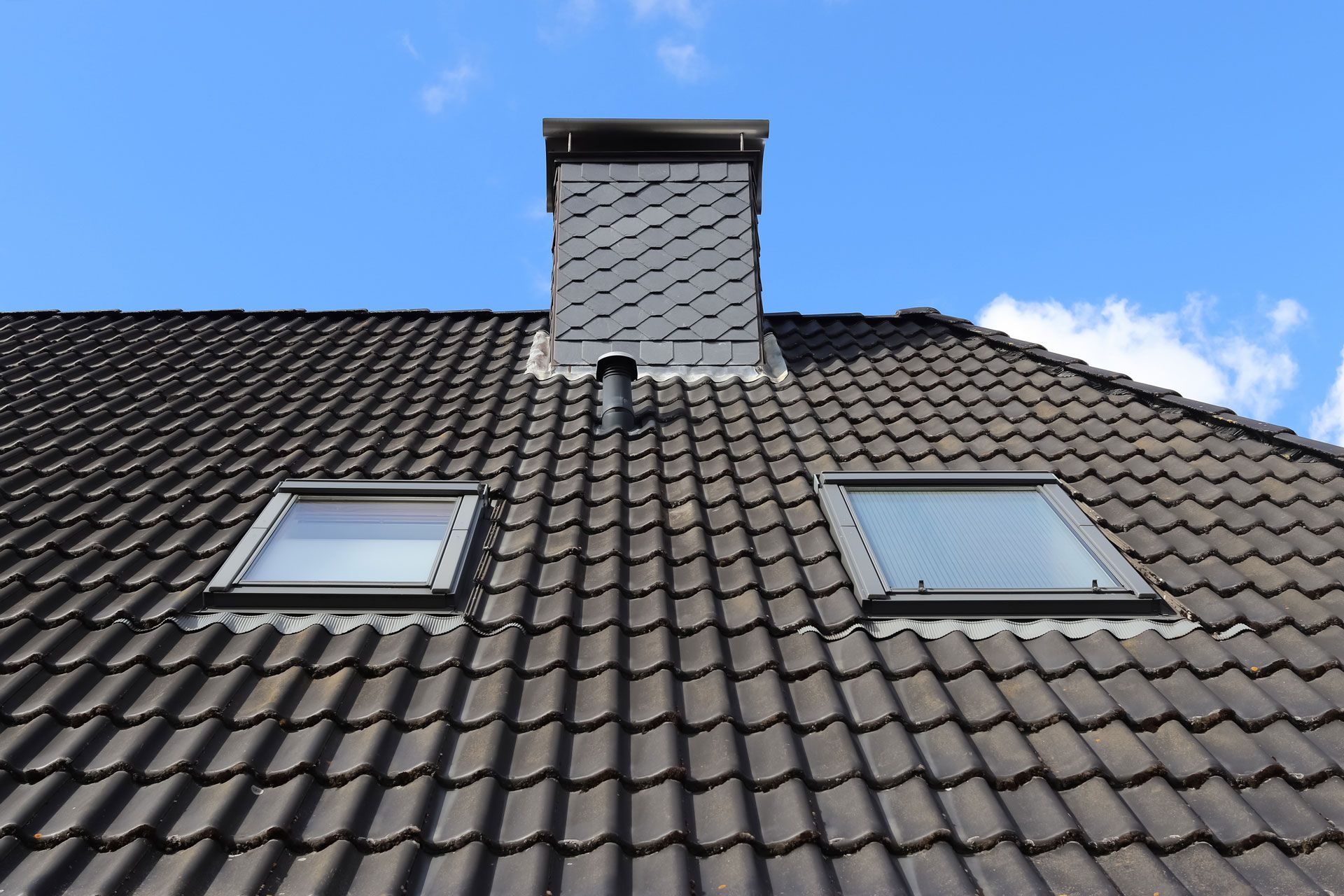 A dark gray tiled roof with two skylights and a chimney against a blue sky.