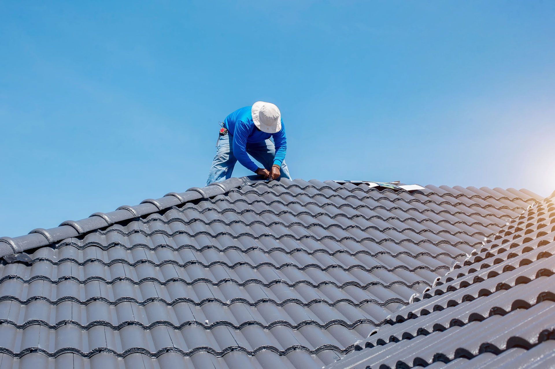 Roofer in blue uniform works on a gray tiled roof under a bright blue sky.