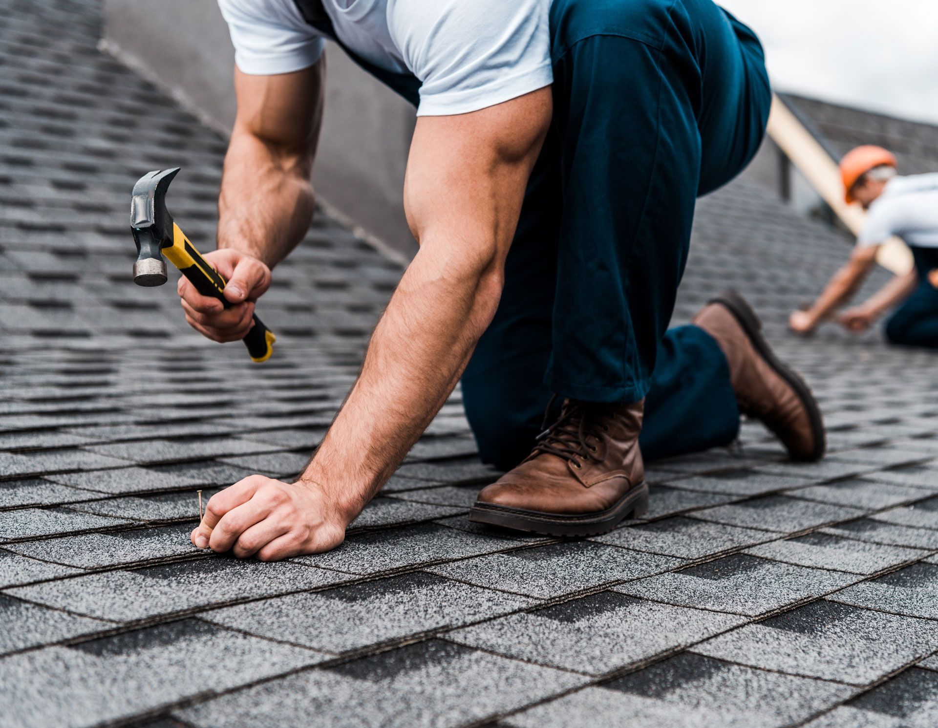 Roofer kneeling, hammering a nail into a gray shingle roof. Another worker in the background.