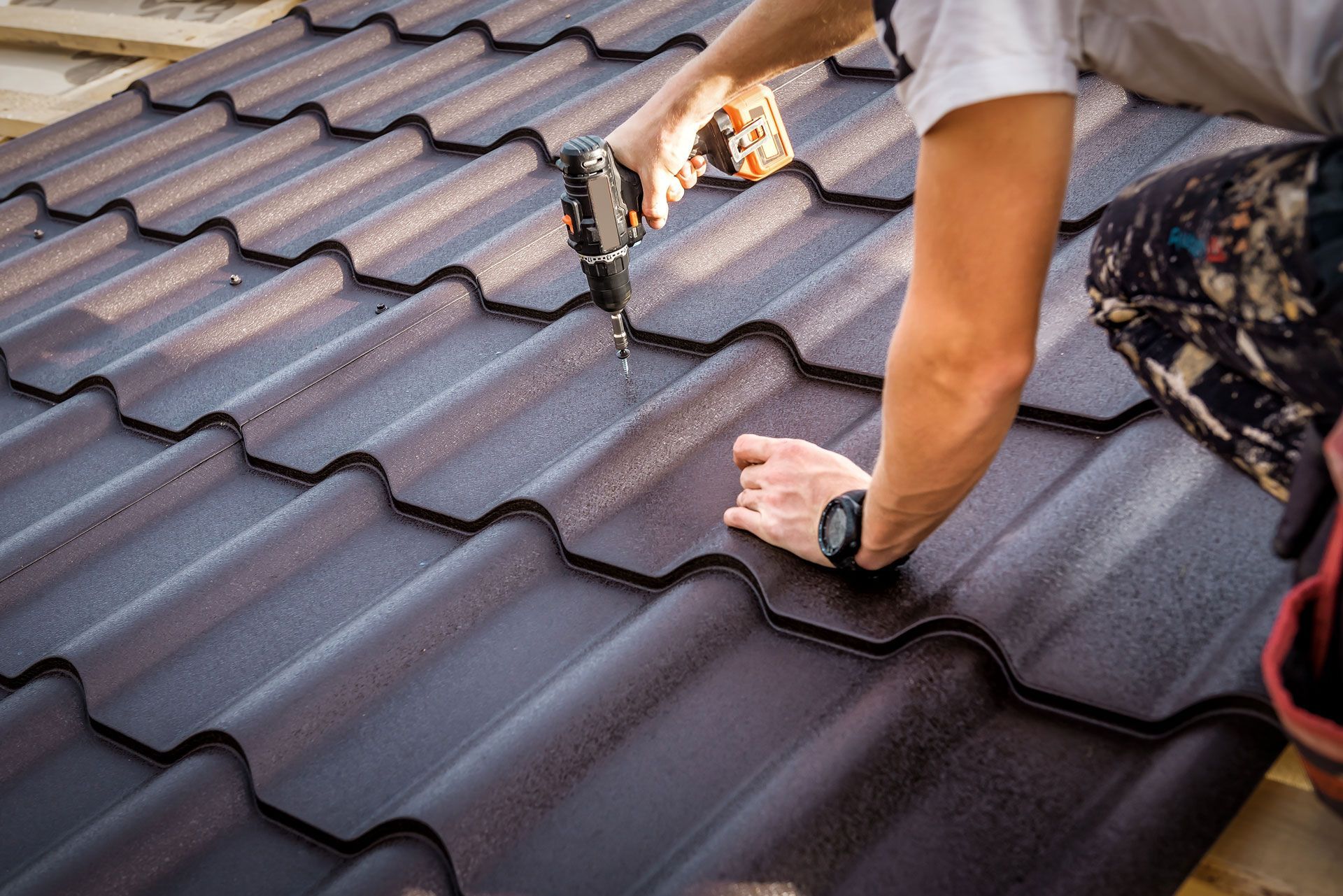 Person installing brown metal roofing with a drill on a wooden roof structure.