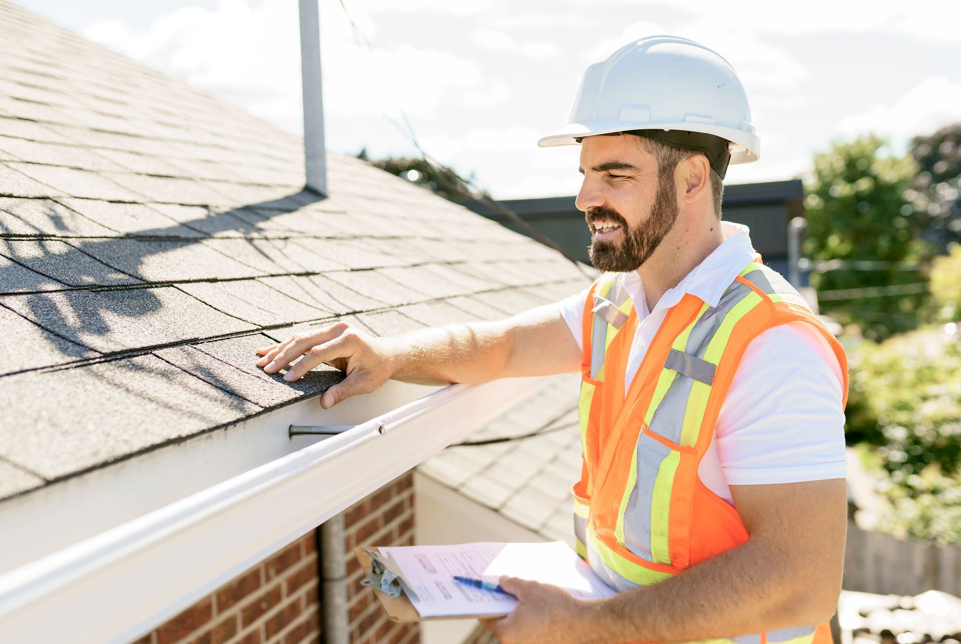 Inspector in hard hat and safety vest inspecting a rooftop gutter; holding a clipboard.