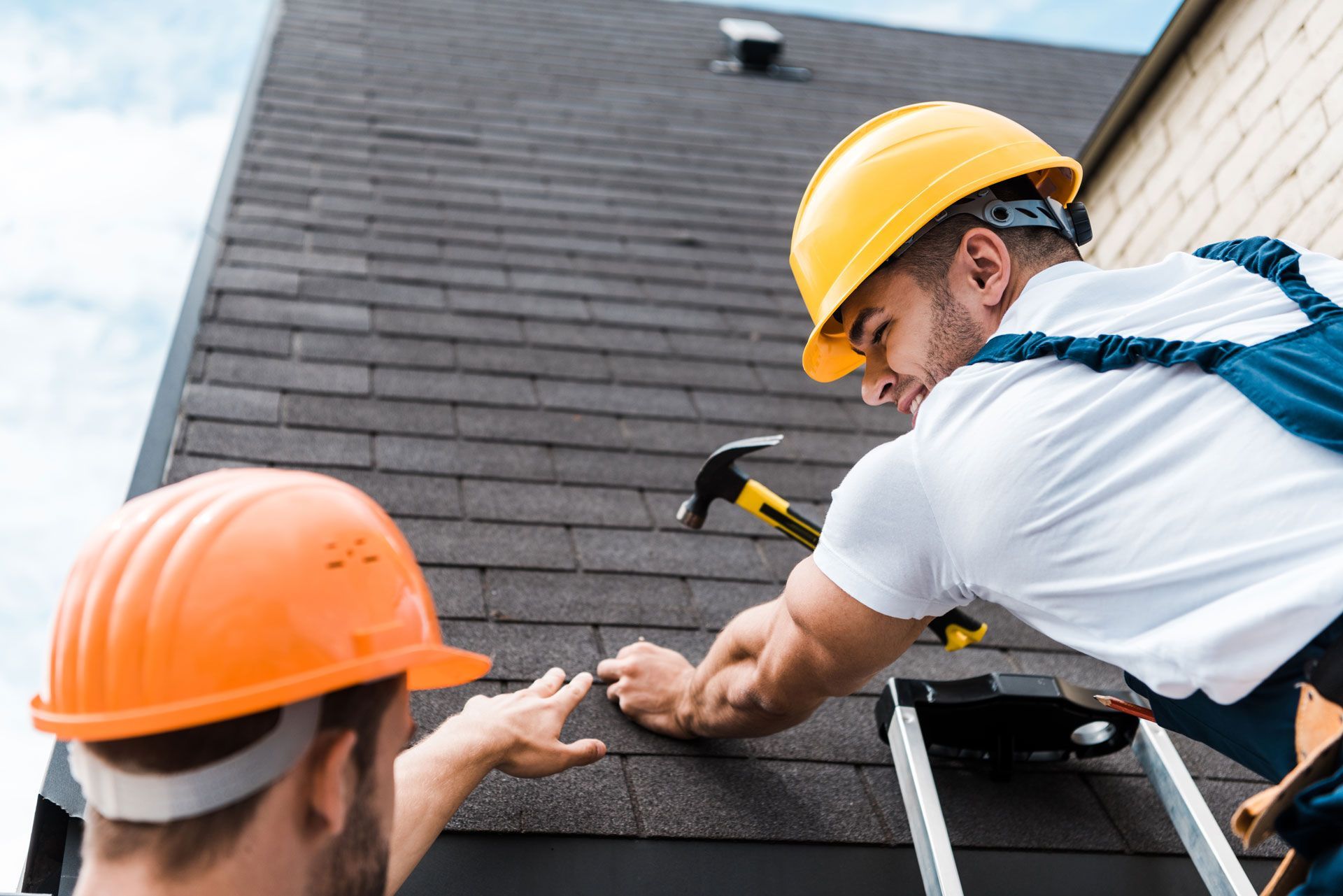Two roofers in hard hats on a rooftop installing shingles with a hammer.