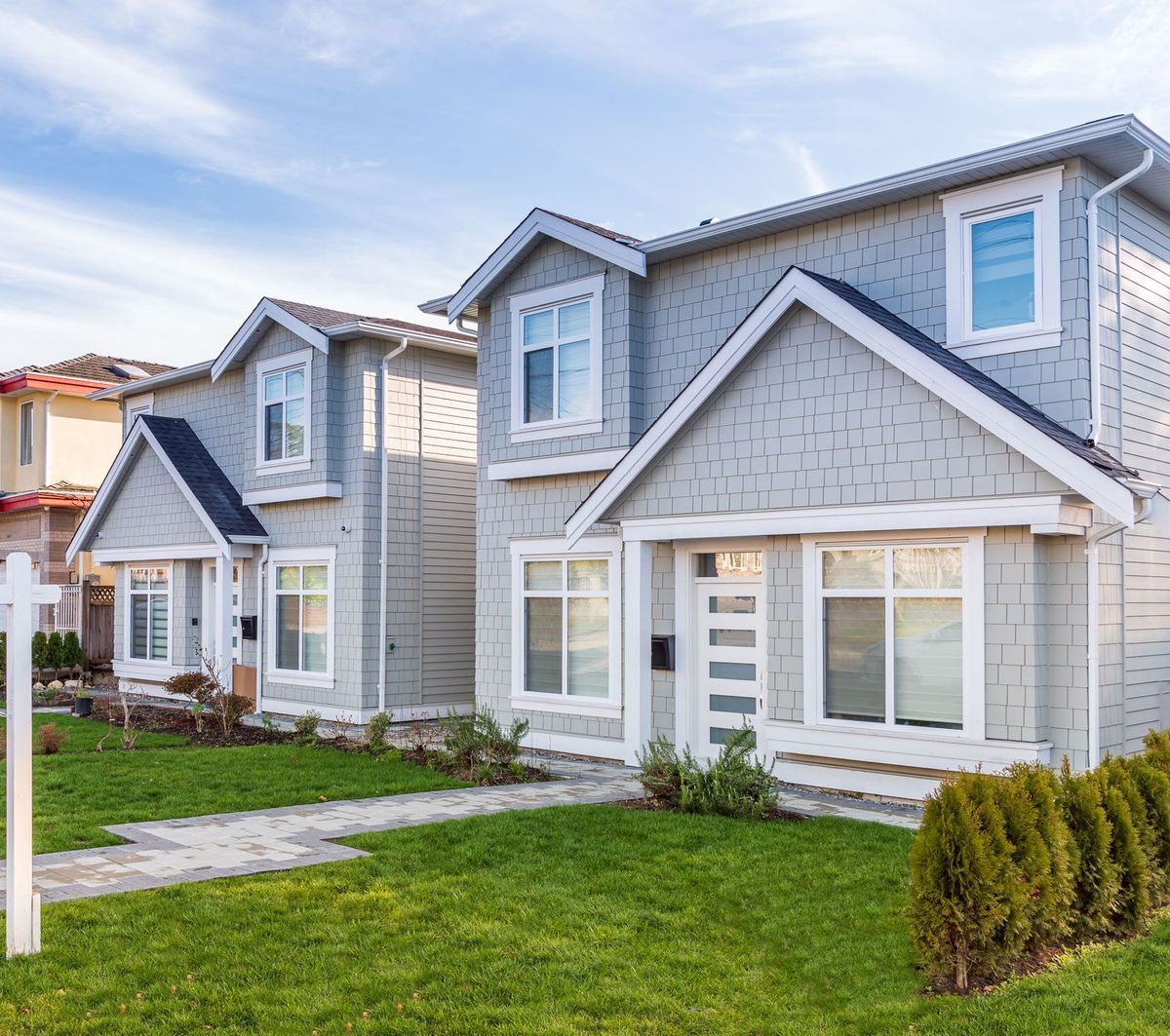 Two gray houses with white trim, a green lawn, and a blue sky.