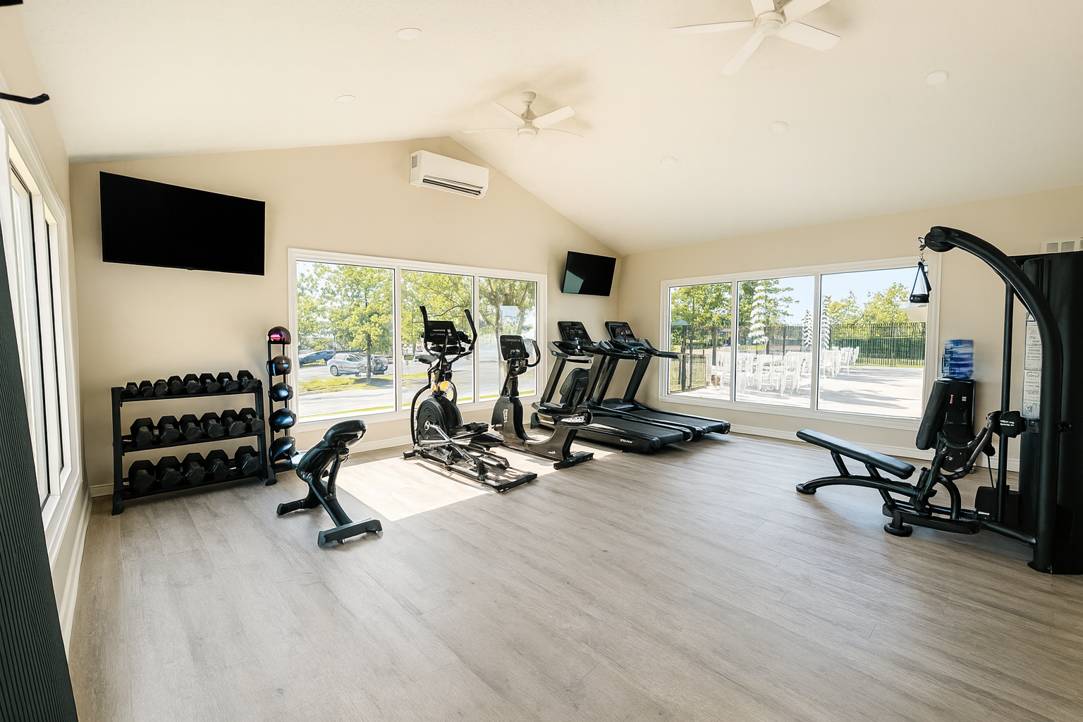 Gym with exercise equipment: weights, treadmills, elliptical, bench. Large windows, light wood floors, TVs.