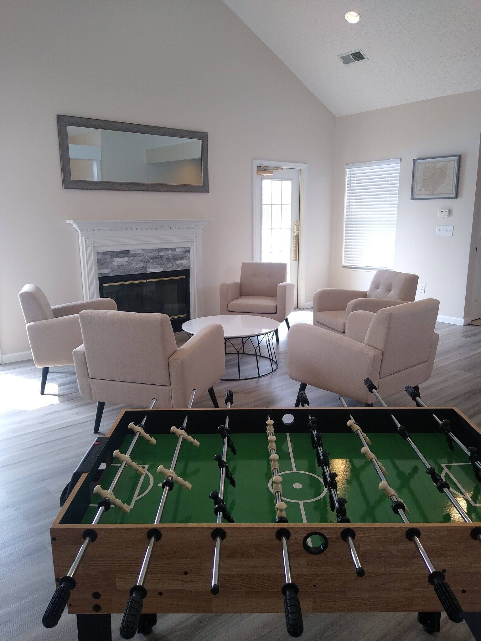 Foosball table in a lounge area with armchairs around a coffee table, fireplace, and white shutters.