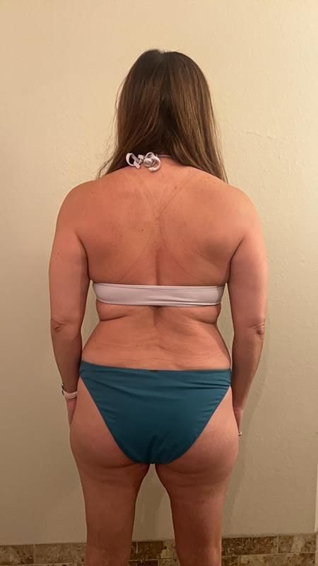 Woman in a bikini from the back, indoors. Light skin, brown hair, wearing a white top and teal bottoms.