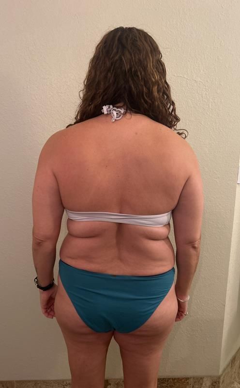 Person in a teal bikini stands facing away from the camera. Tan skin, curly hair, against a neutral wall.