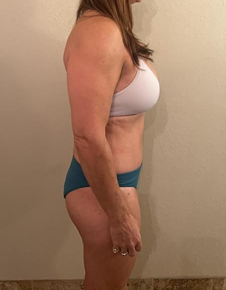 Woman in white bra and teal underwear stands sideways against a wall.