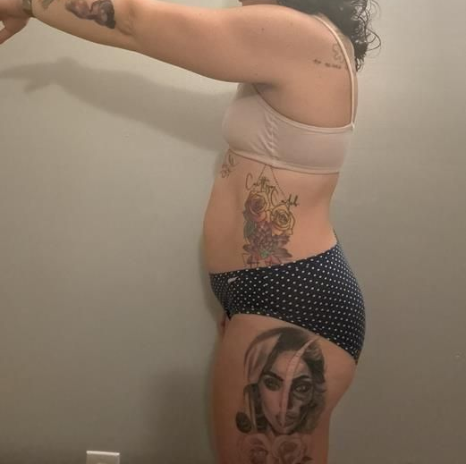 Woman in underwear with tattoos, standing sideways against a plain wall.