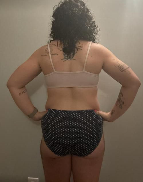 Person in a beige bra and polka dot underwear with arms at their sides, standing in front of a gray wall.