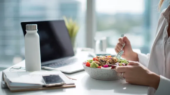 Person eating a salad at a desk with a laptop, water bottle, and notebook.
