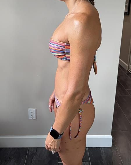 Woman in a strapless bikini stands in front of a neutral wall. Her arms are flexing, and she's wearing a watch.